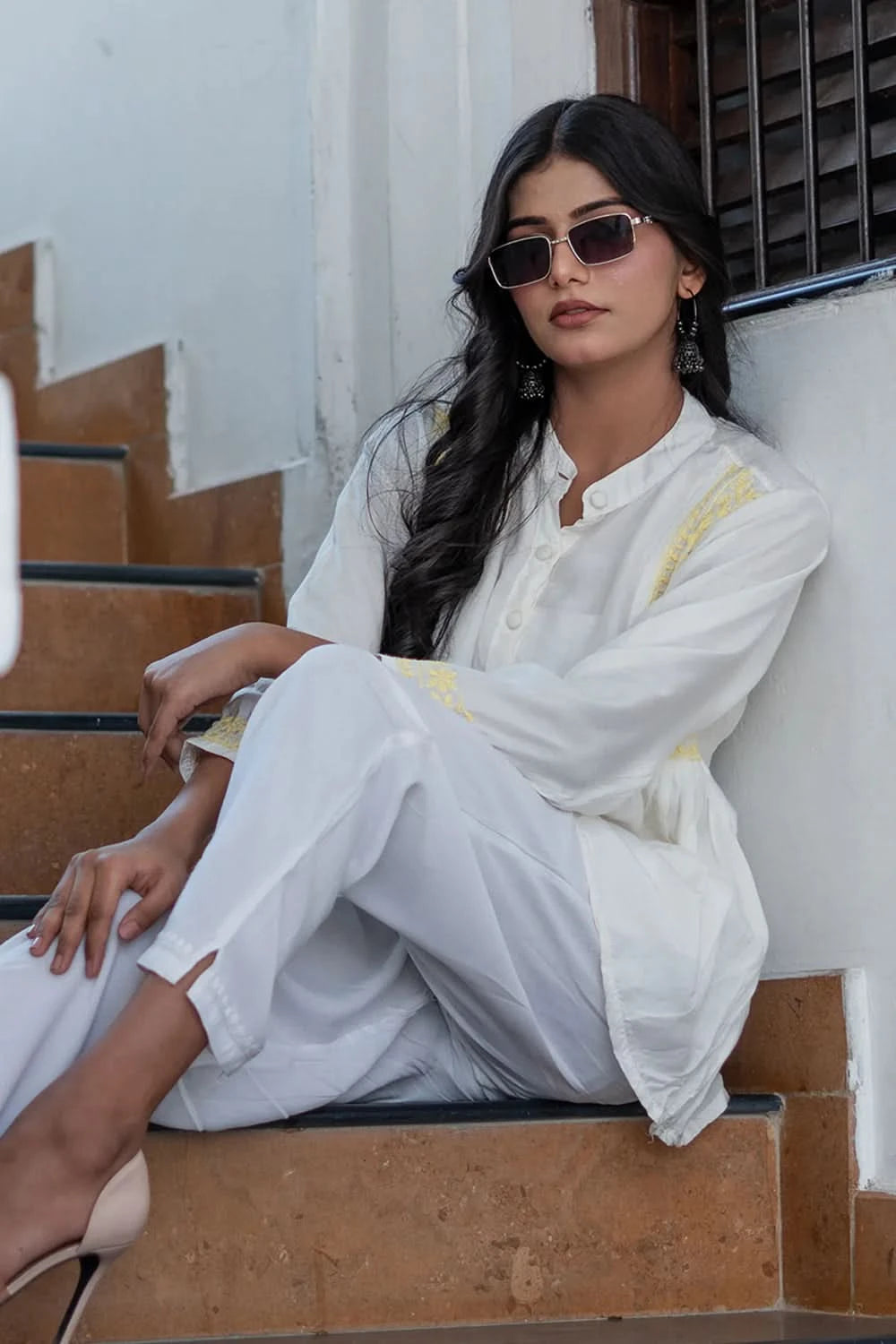 Woman in a white outfit sitting on steps wearing sunglasses.