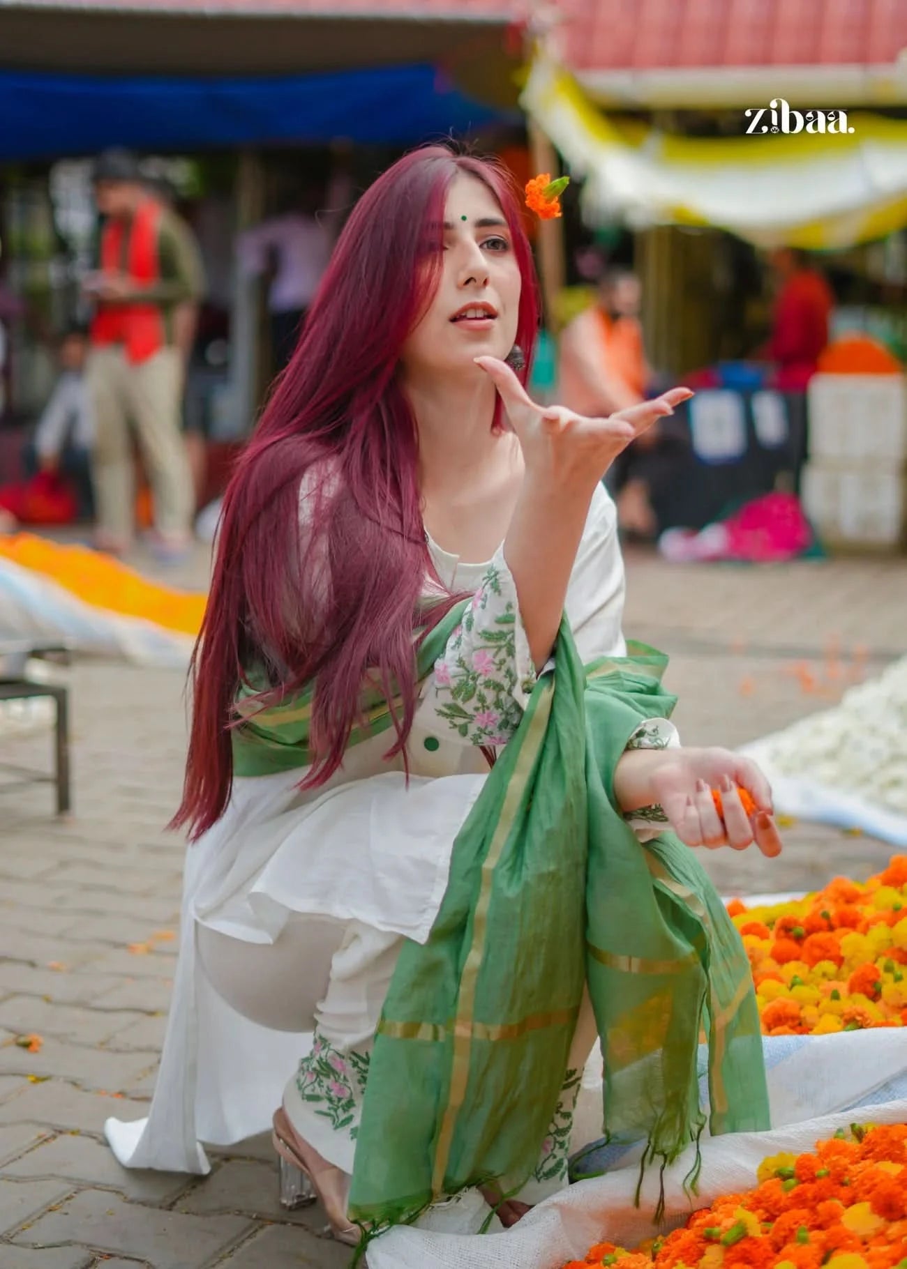 Woman with red hair and green scarf sitting among flowers at a market.