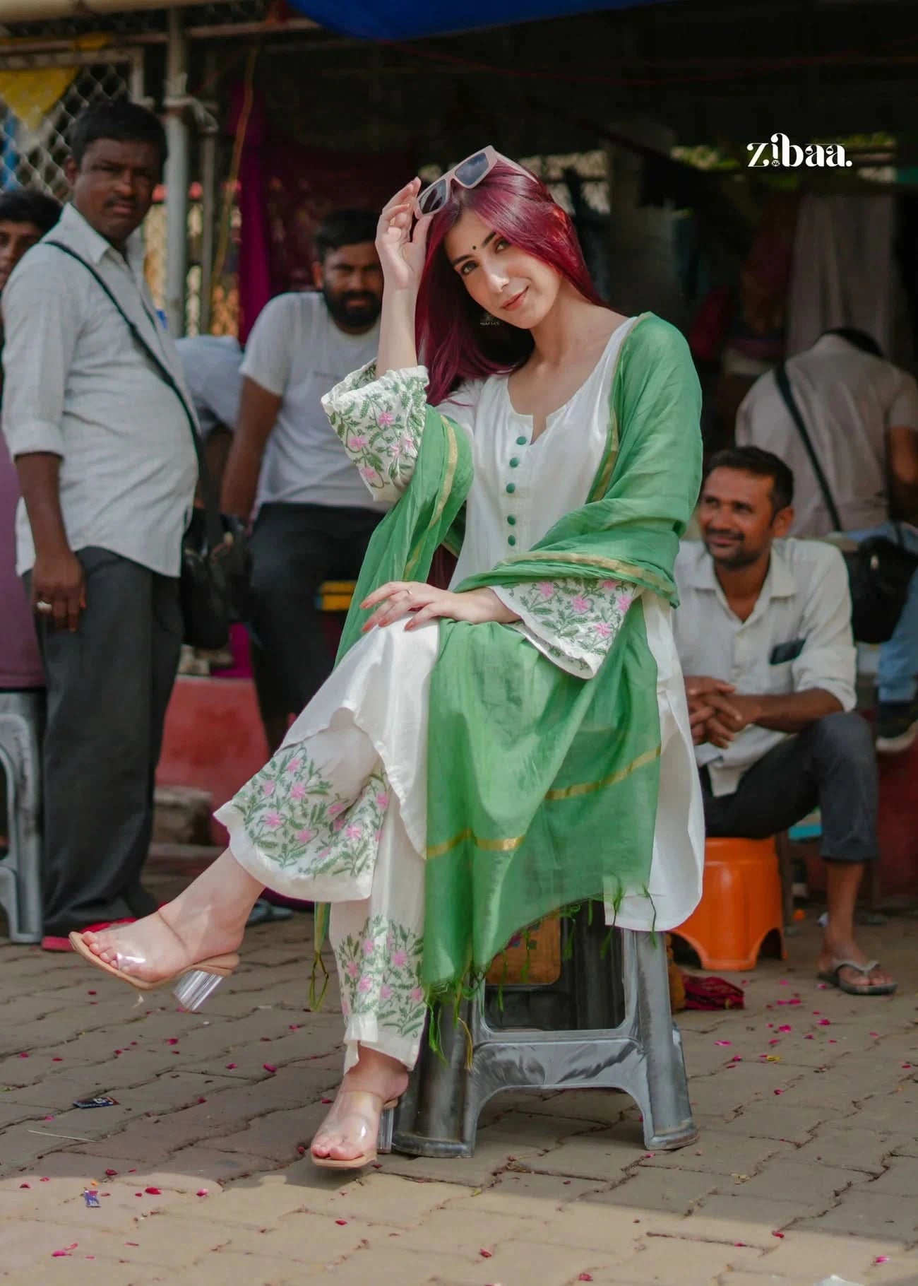 Woman in a green and white traditional outfit sitting on a stool in an outdoor setting.