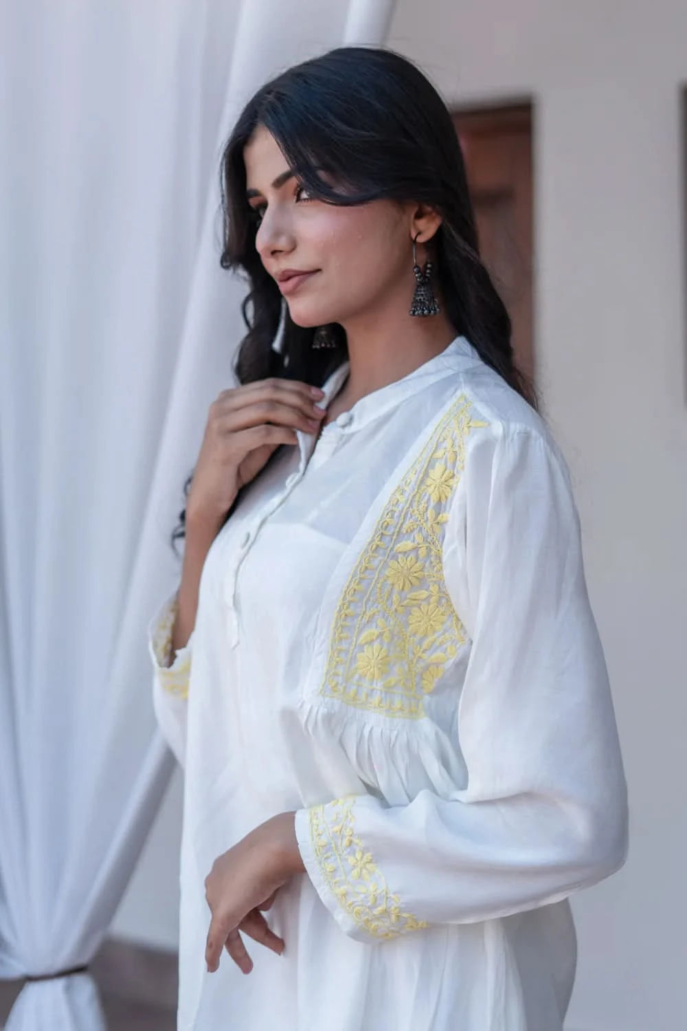 Woman wearing a white embroidered blouse with yellow details, standing against a white curtain.