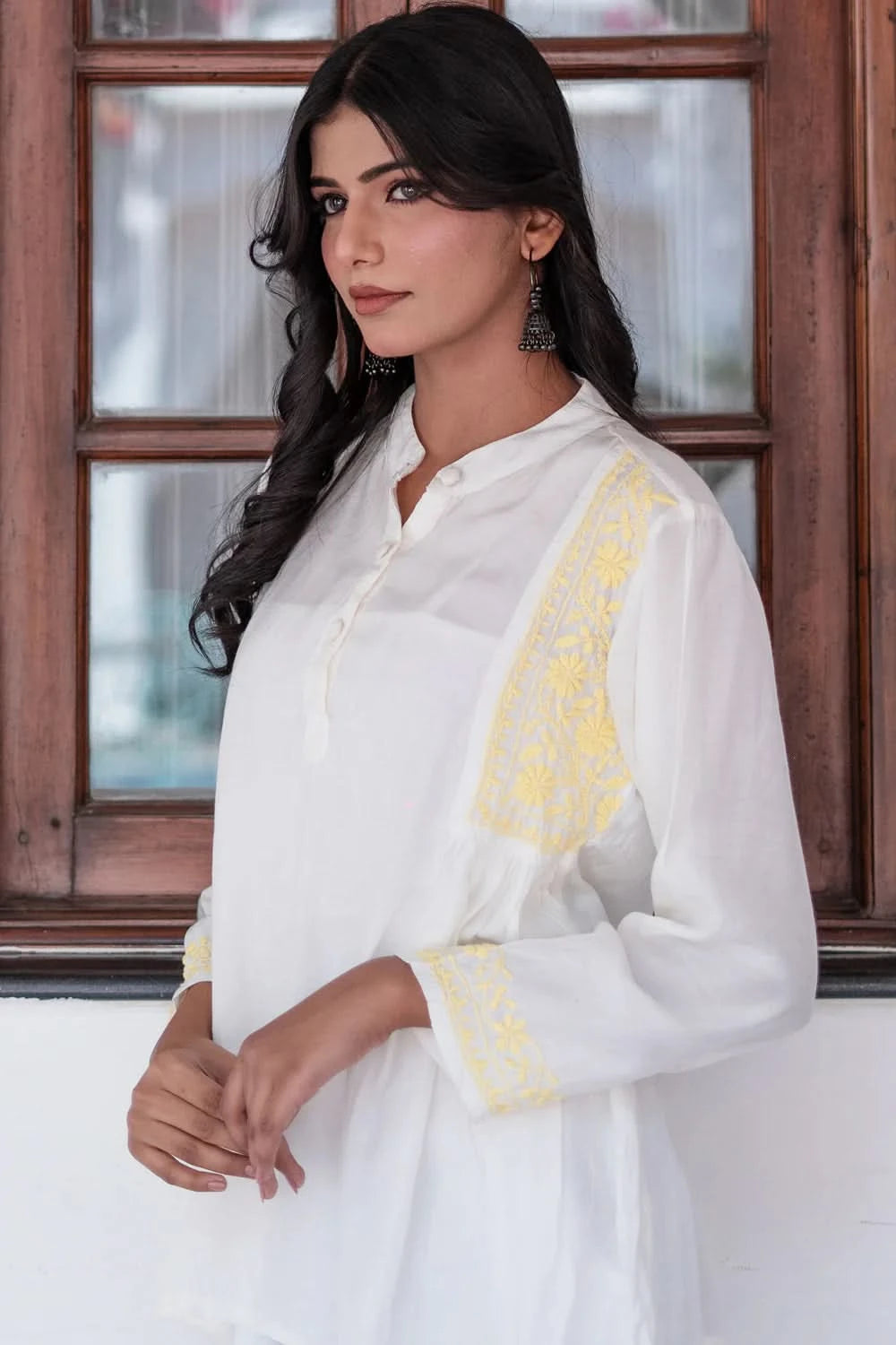 Woman wearing a white outfit with yellow accents in front of a wooden window.