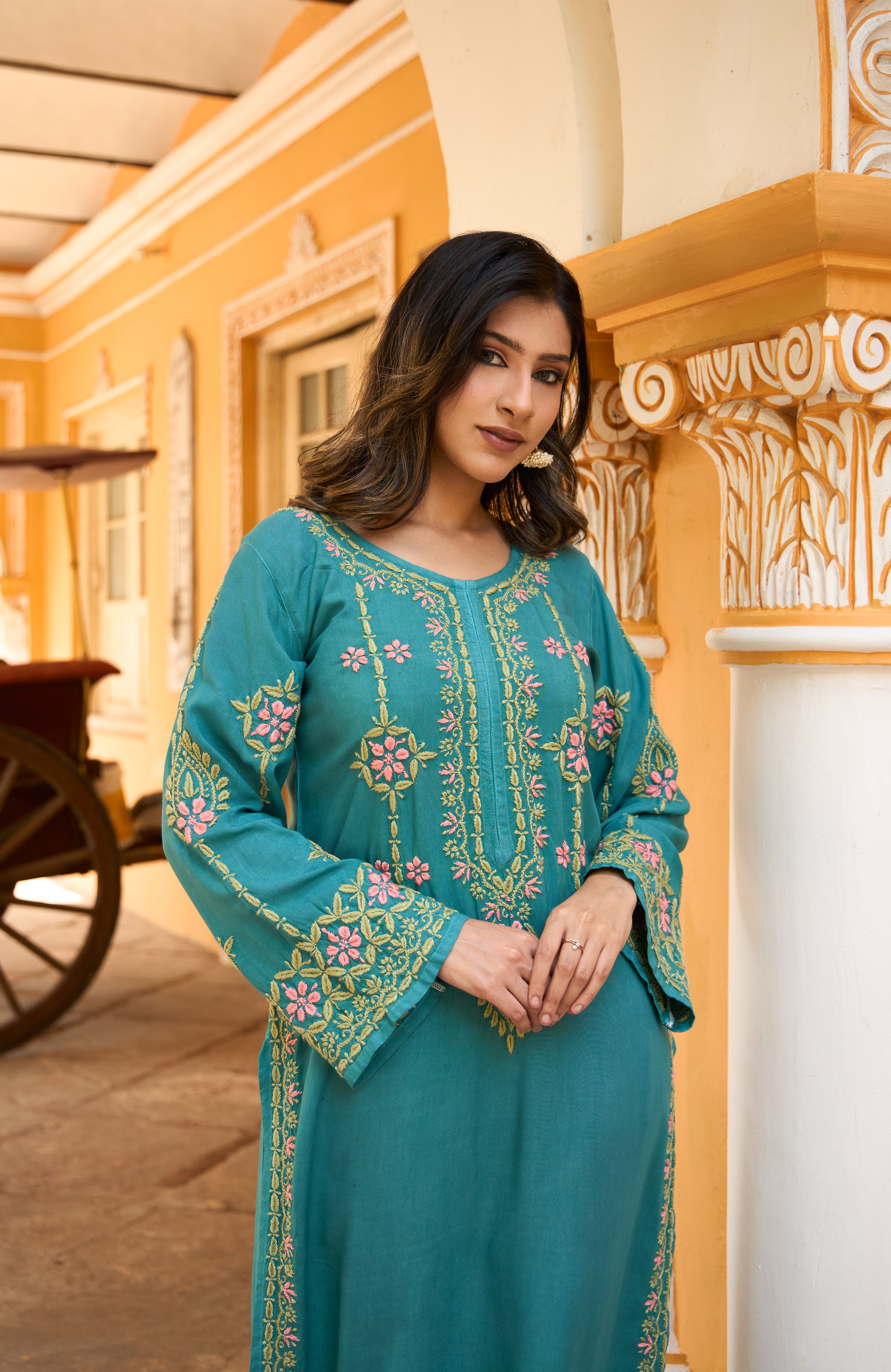 Woman in a teal embroidered outfit standing in front of a yellow building.