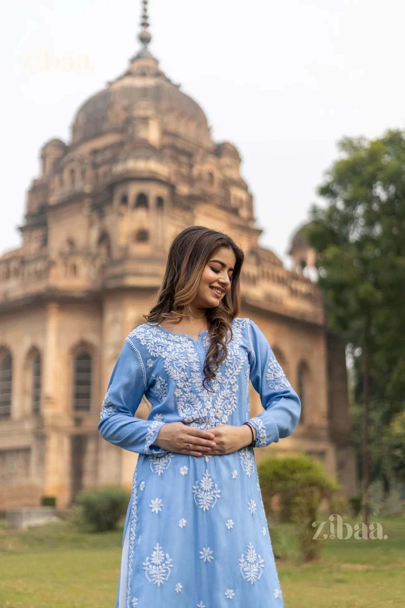 The model in a Blue Chikankari Kurta with white embroidery poses gracefully at the base of the historic monument in a lush green area.