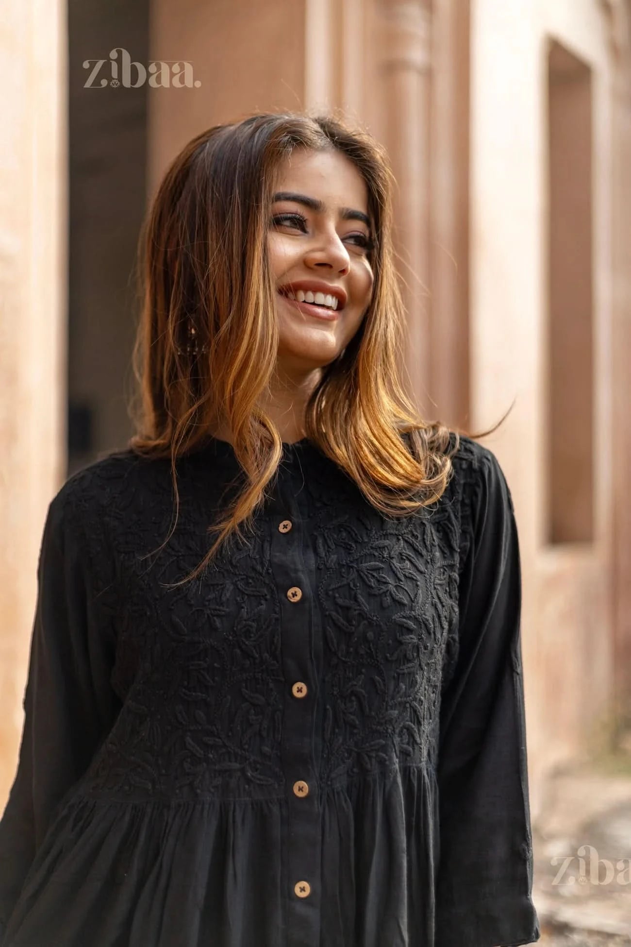 A woman smiles in an outdoor setting, wearing a black Chikankari top with detailed embroidery and button accents, exuding elegance.