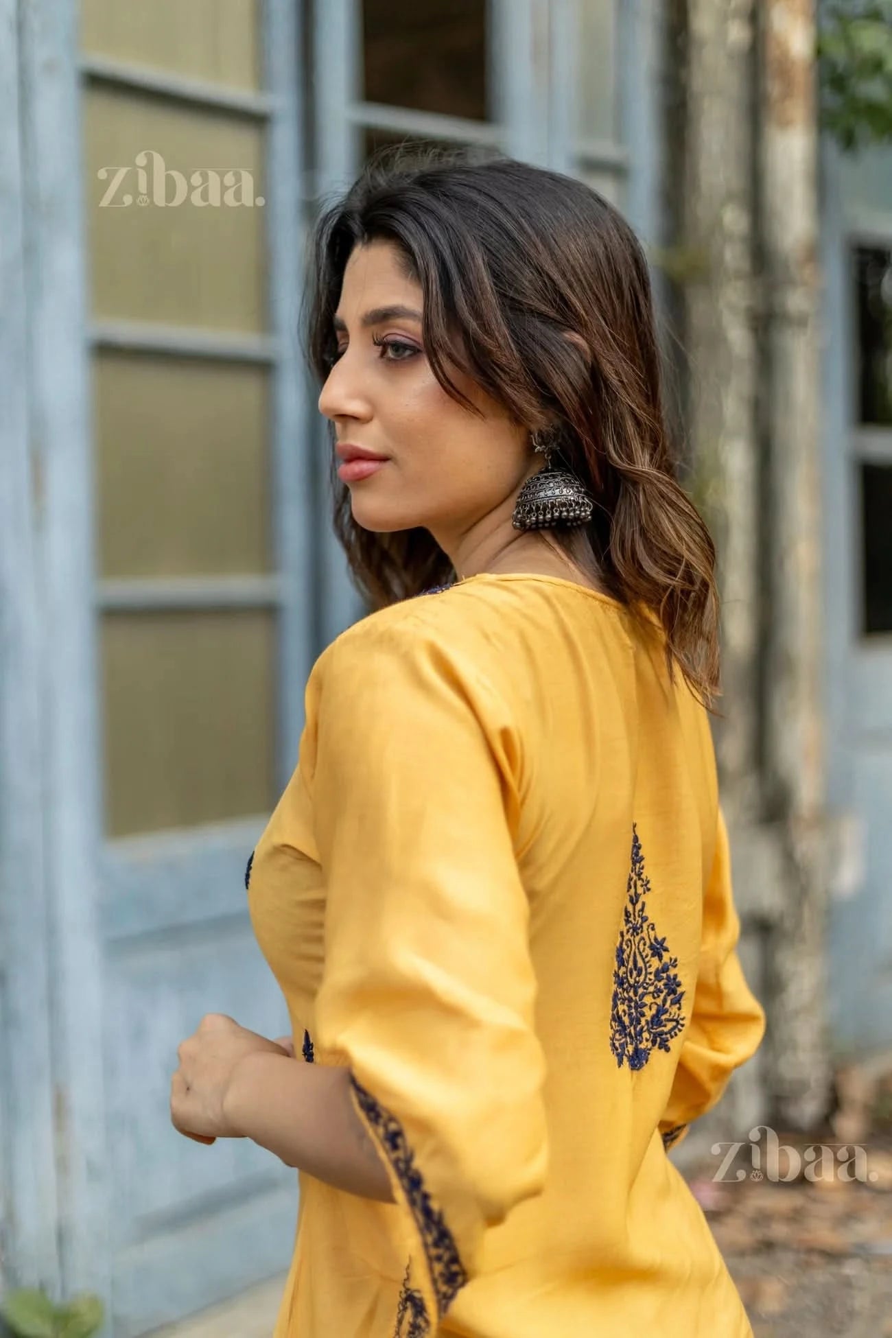 A woman poses in a mustard Chikankari Kurta Set, showing intricate navy embroidery on the back and sleeves, paired with earrings.