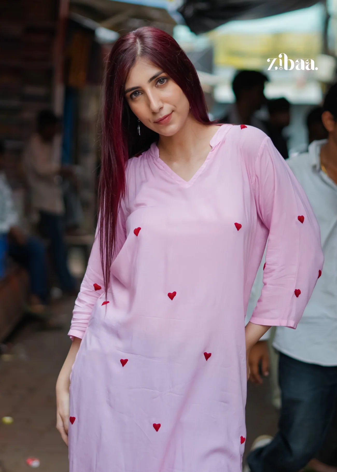 A woman stands on a bustling street wearing a pink chikankari kurta, featuring delicate red embroidered heart motifs.