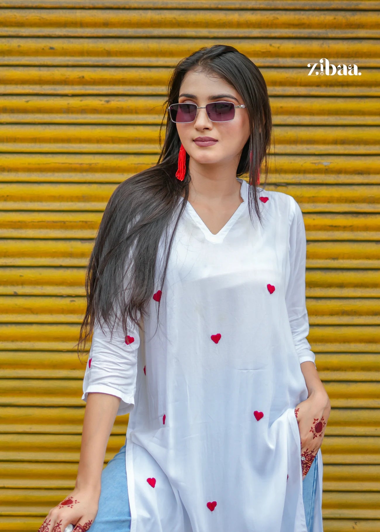 The model poses casually in front of a bright yellow shop shutter, wearing a white chikankari kurti decorated with red heart motifs.