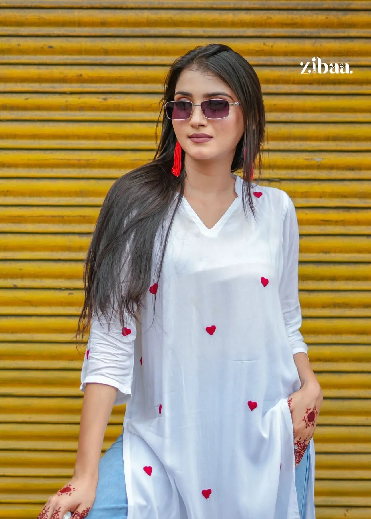 The model poses casually in front of a bright yellow shop shutter, wearing a white chikankari kurti decorated with red heart motifs.