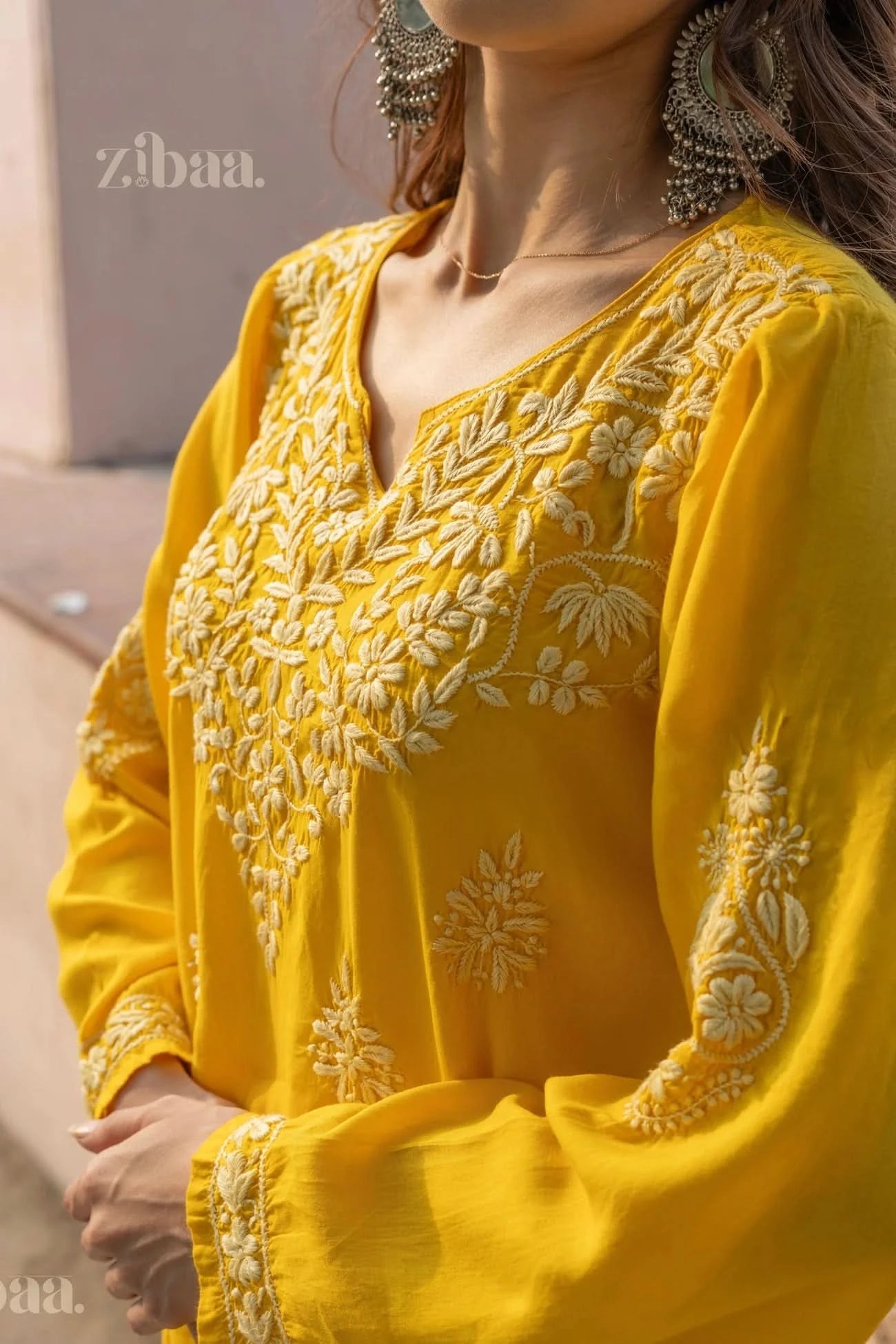 Close-up of a woman wearing a yellow Chikankari kurti with detailed floral embroidery, arms crossed lightly, posing outdoors in sunlight.