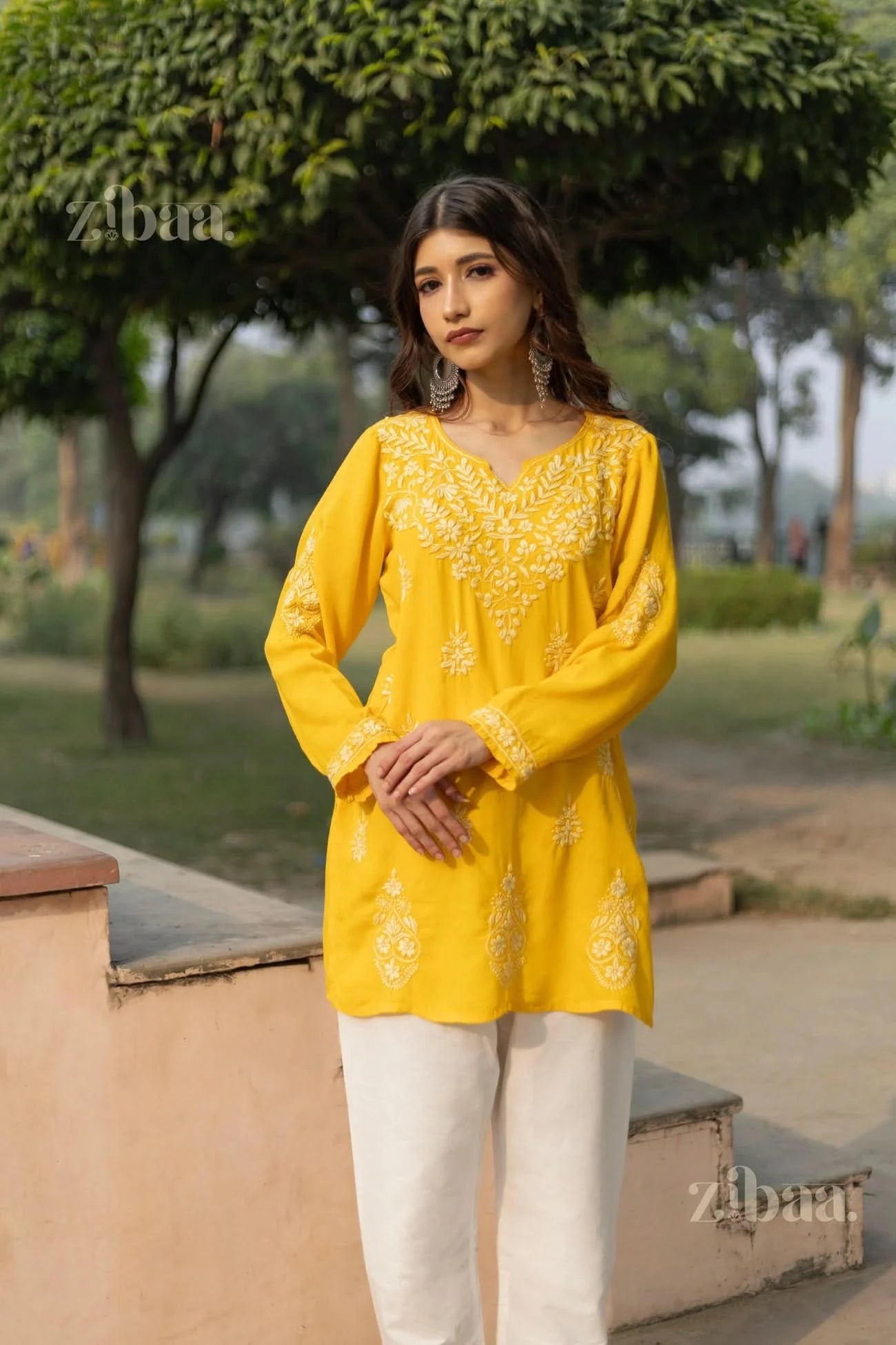 A woman wearing a yellow short kurti with white floral embroidery stands with hands clasped in front, looking confidently at the camera.