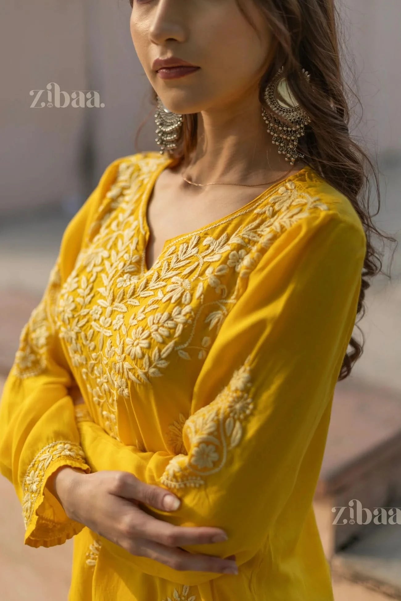 A woman wearing a yellow Chikankari kurti with intricate embroidery crosses her arms softly, standing outdoors and looking to the side.
