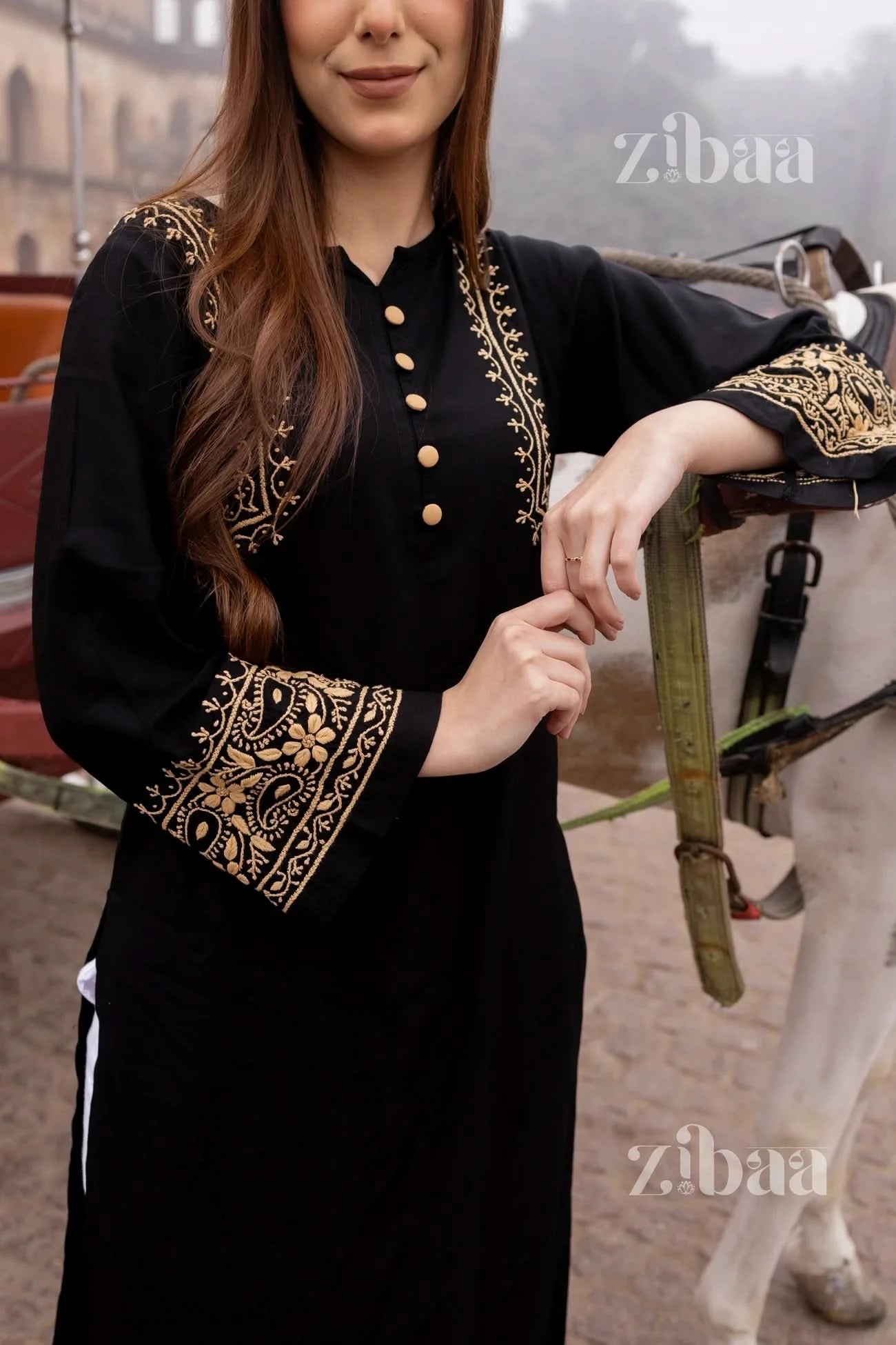 Woman modeling black chikankari kurti with floral embroidery standing outside with a horse cart in the backdrop.
