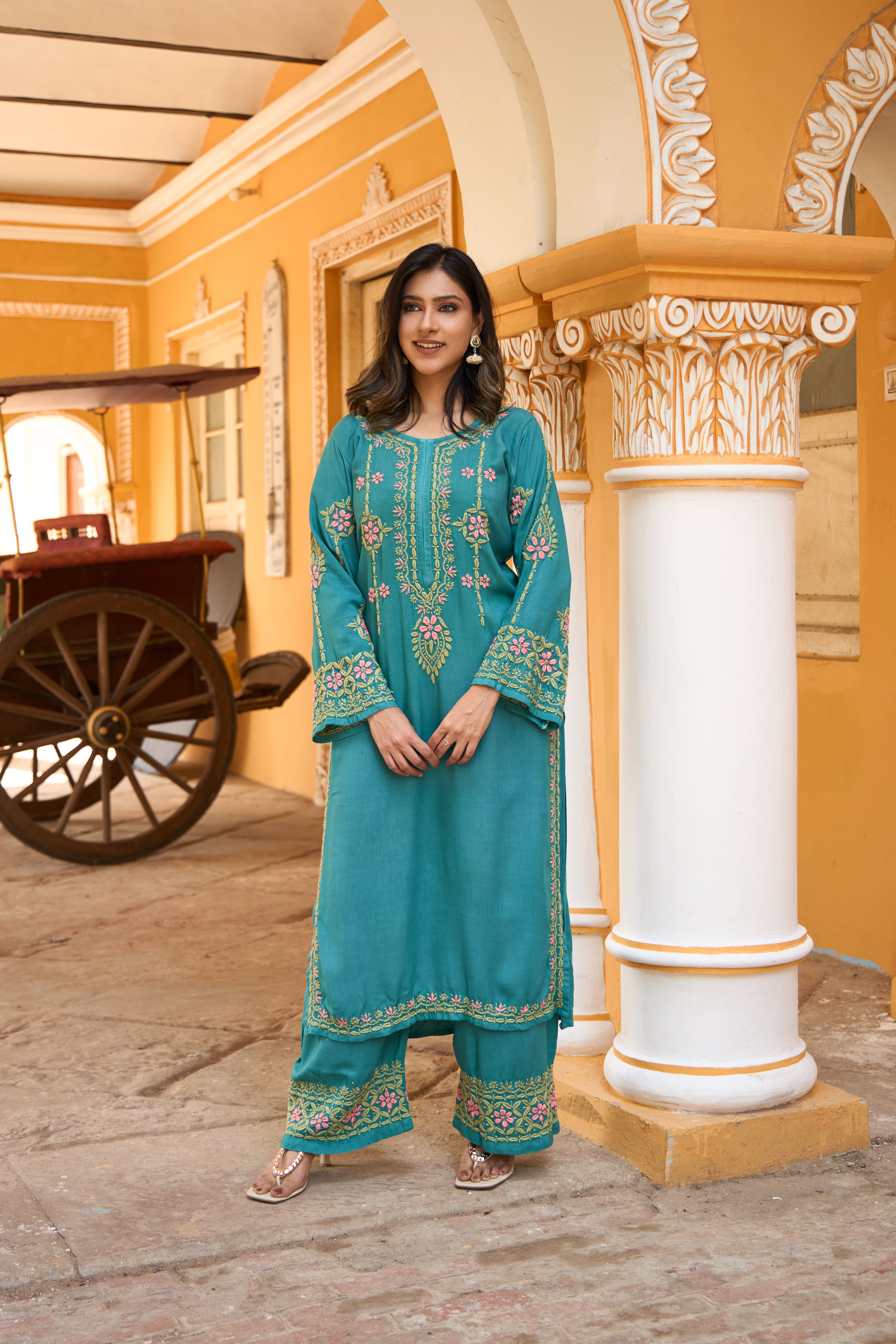Woman in a teal chikankari kurta set standing in front of a decorative building with a cart in the background.