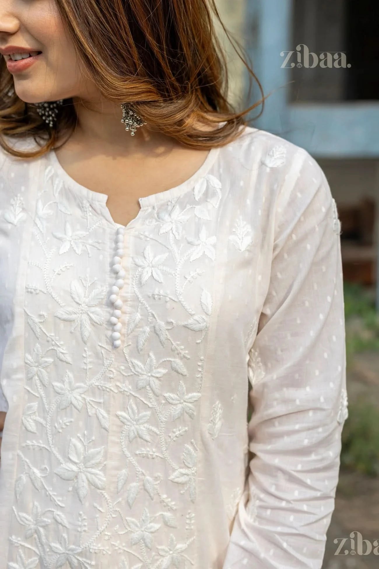 Close-up of a woman wearing a white Chikankari Anarkali with floral embroidery, slightly turned to the side, smiling softly outdoors.