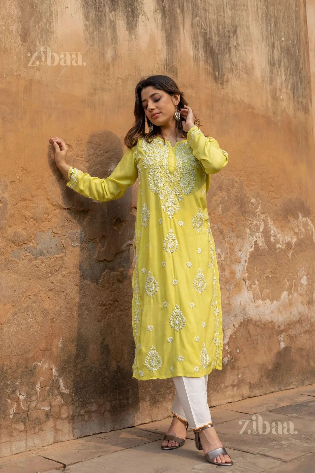 A woman leans against a textured wall in a lime green Chikankari Kurti with white embroidery, adjusting her hair, paired with white pants.