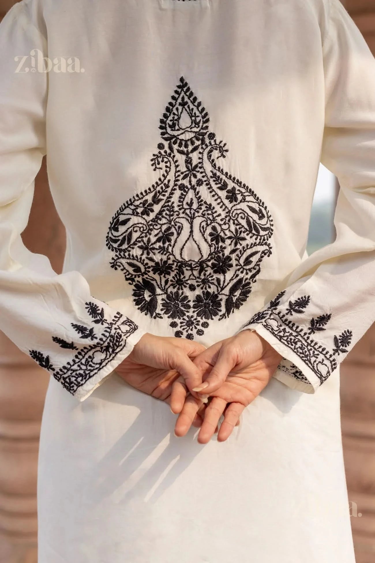 Back view of a white Chikankari kurta set with detailed black embroidery, as the woman clasps her hands behind her, standing outdoors.