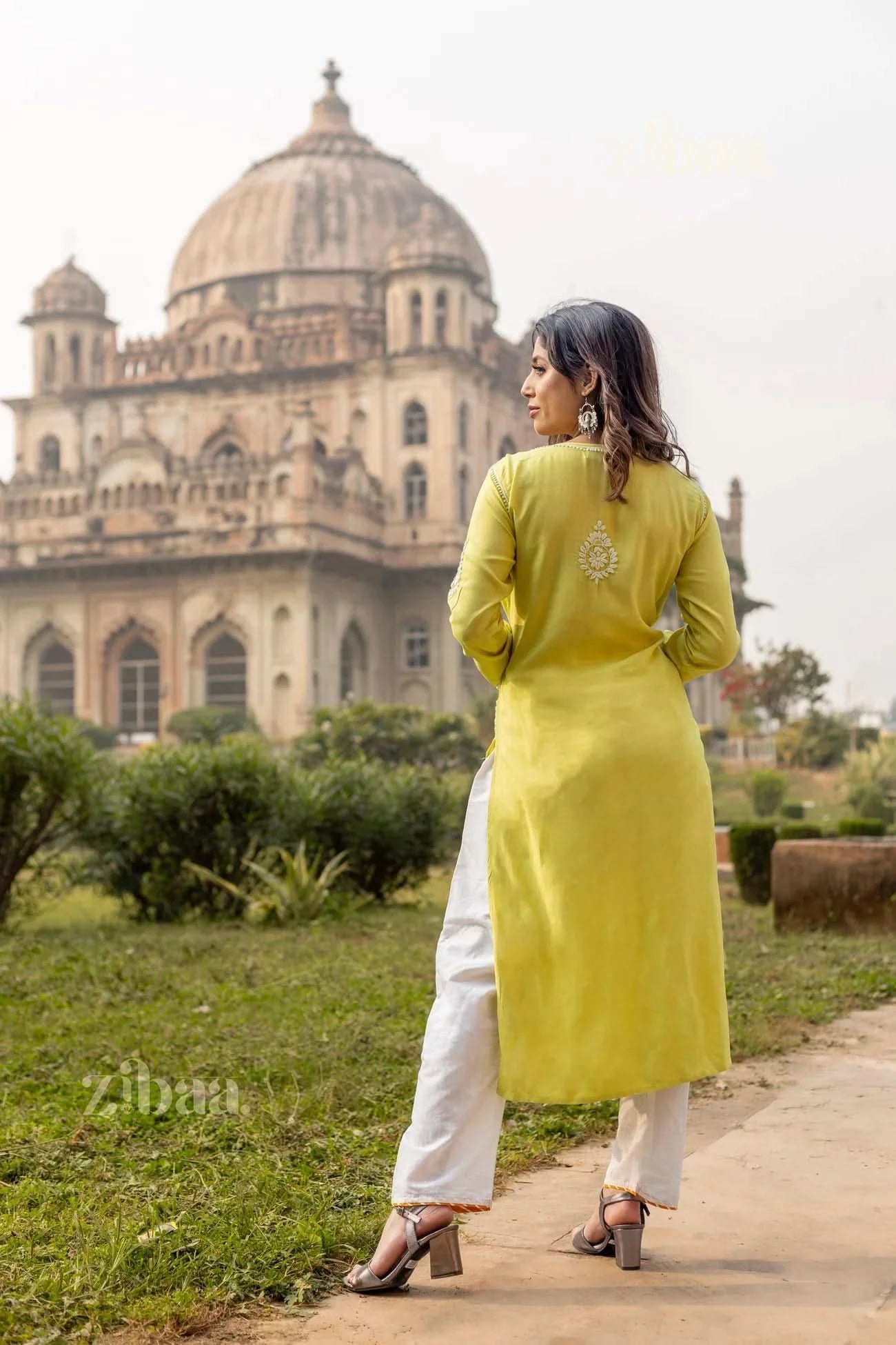A woman stands outdoors with her back to the camera, wearing a lime green Chikankari long kurti,, gazing at a historic monument.