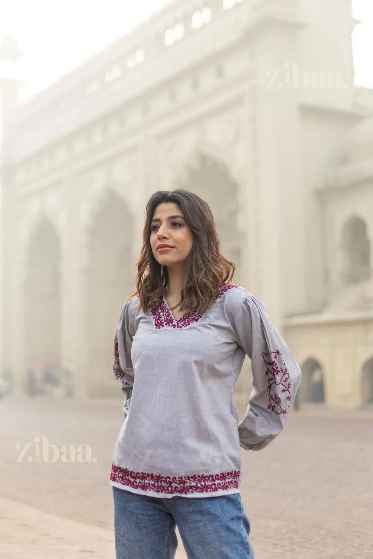 Woman posing in front of a heritage structure, wearing a grey Chikankari top with red embroidery on the neckline, sleeves, and hem