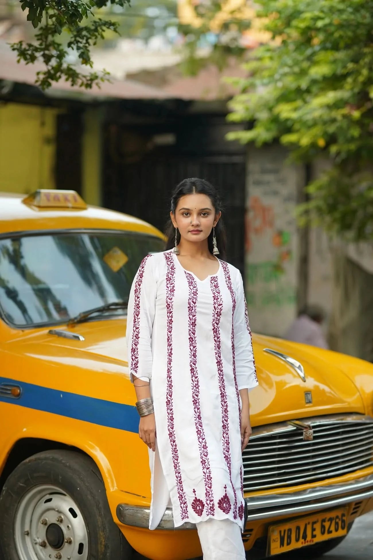 Woman in a white embroidered kurta standing next to a yellow taxi with greenery in the background