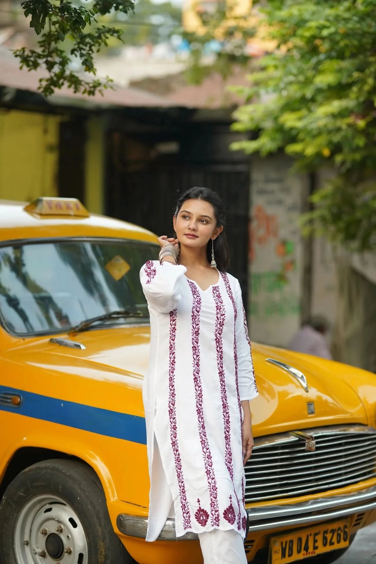 Woman in a white embroidered outfit standing next to a yellow taxi in an urban setting