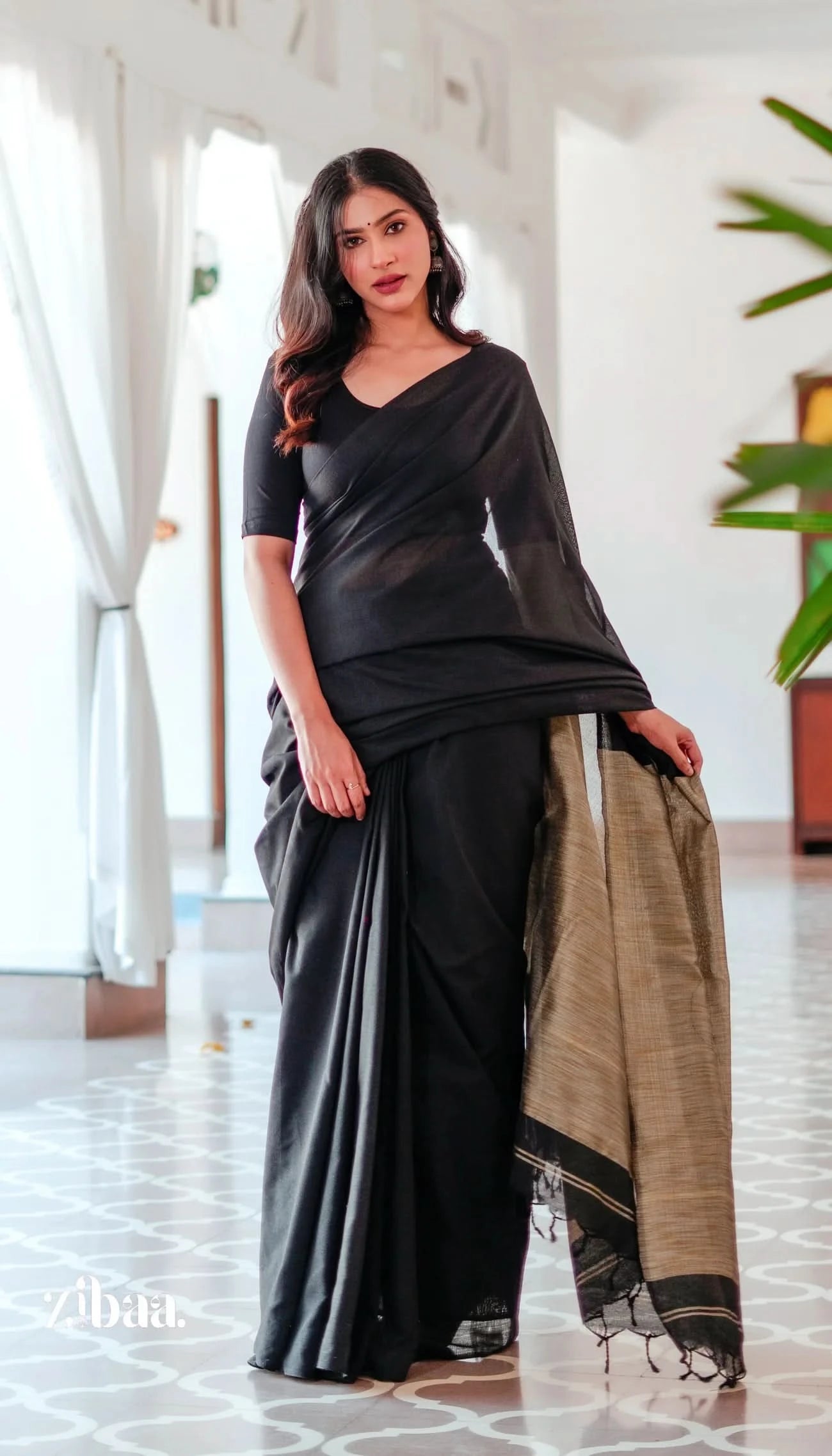 Woman in a black saree with a beige border standing indoors.