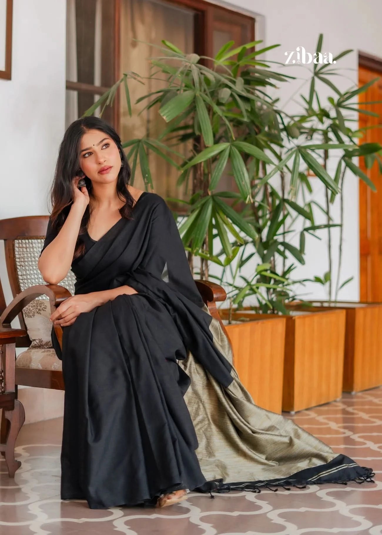 Woman in a black saree sitting indoors with plants in the background