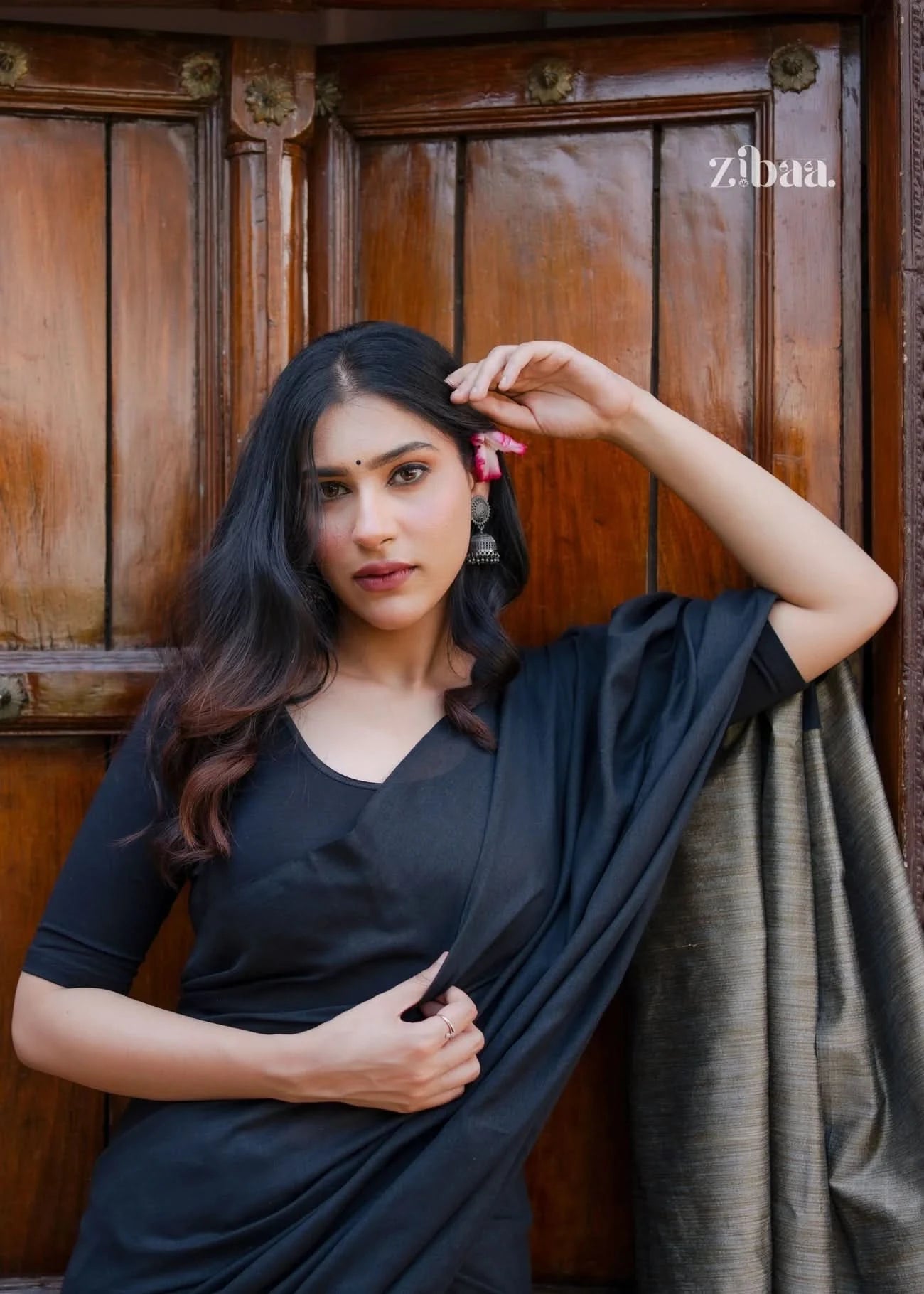 Woman in a black saree standing against a wooden door, with 'zibaa!' branding.