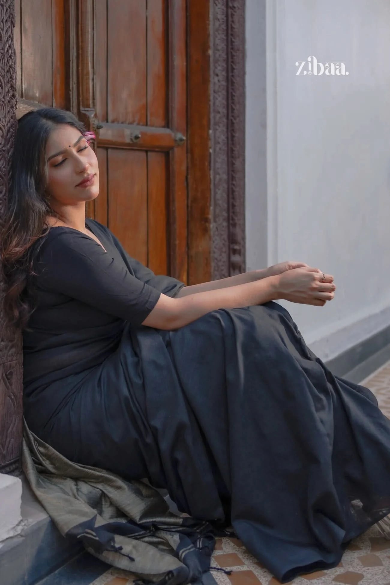 Woman in a dark blue saree sitting against a wooden door.