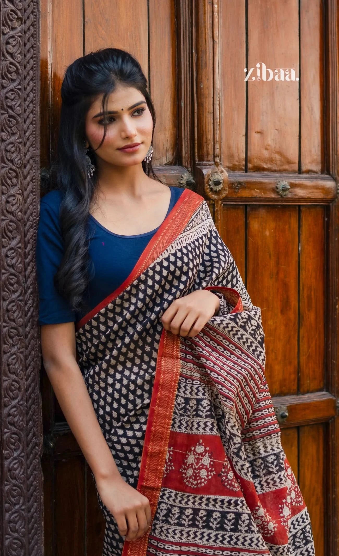 Woman wearing a patterned saree in front of a wooden door with 'zibaa' branding.