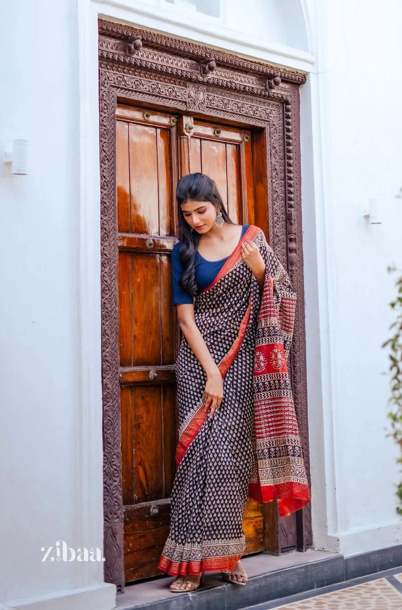 Woman in a saree standing in front of a wooden door with intricate carvings.