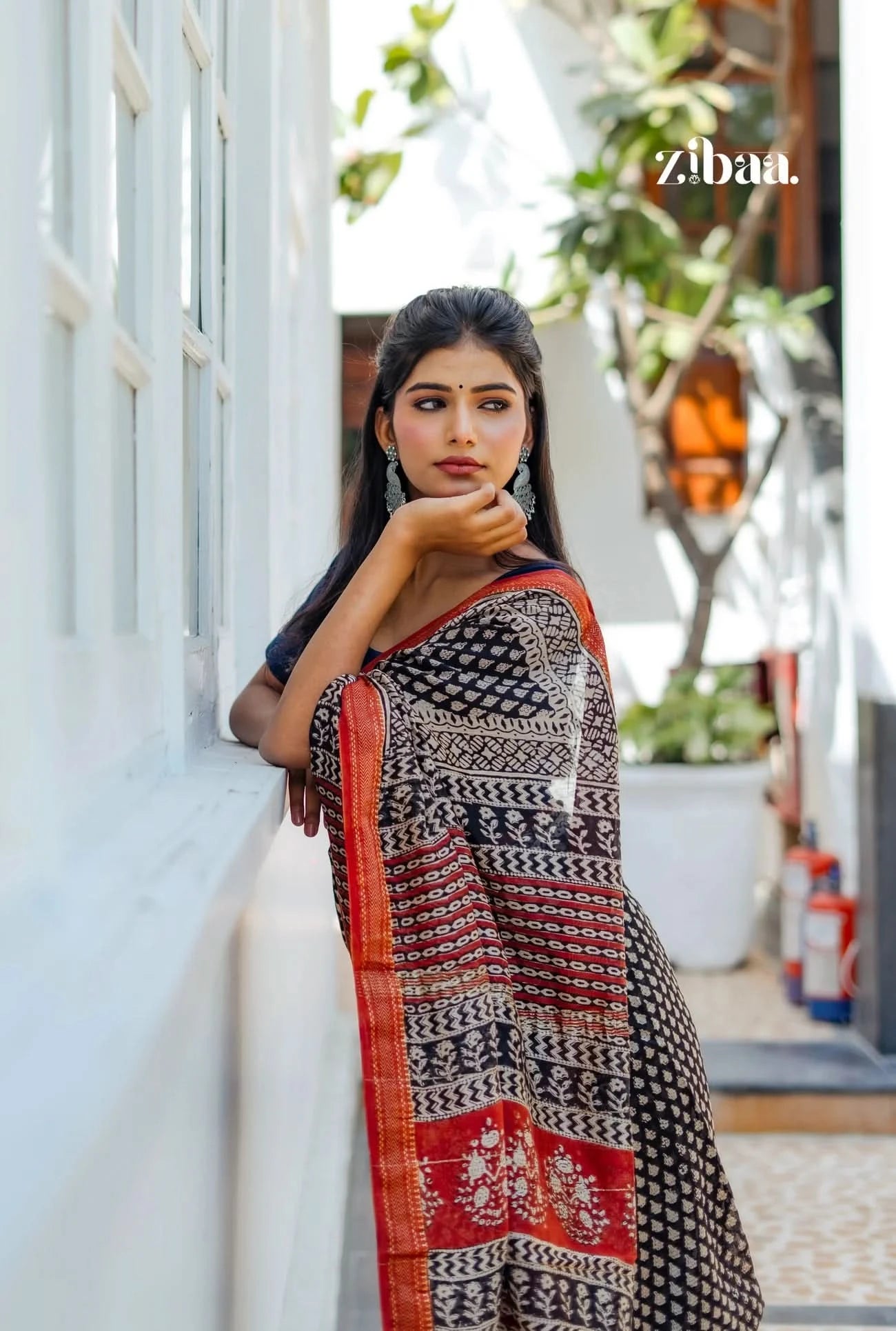 Woman wearing a traditional saree leaning against a white wall with plants in the background