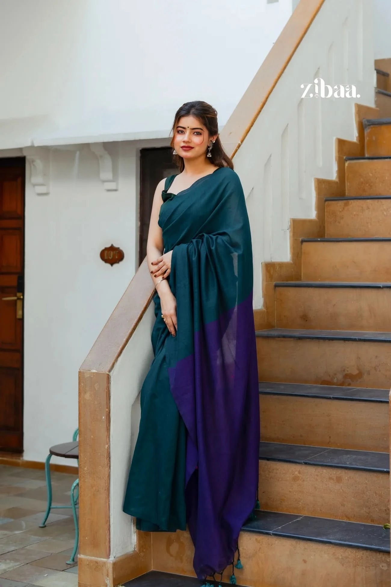 Woman in a teal and purple saree standing on a staircase with 'zibaa' branding.