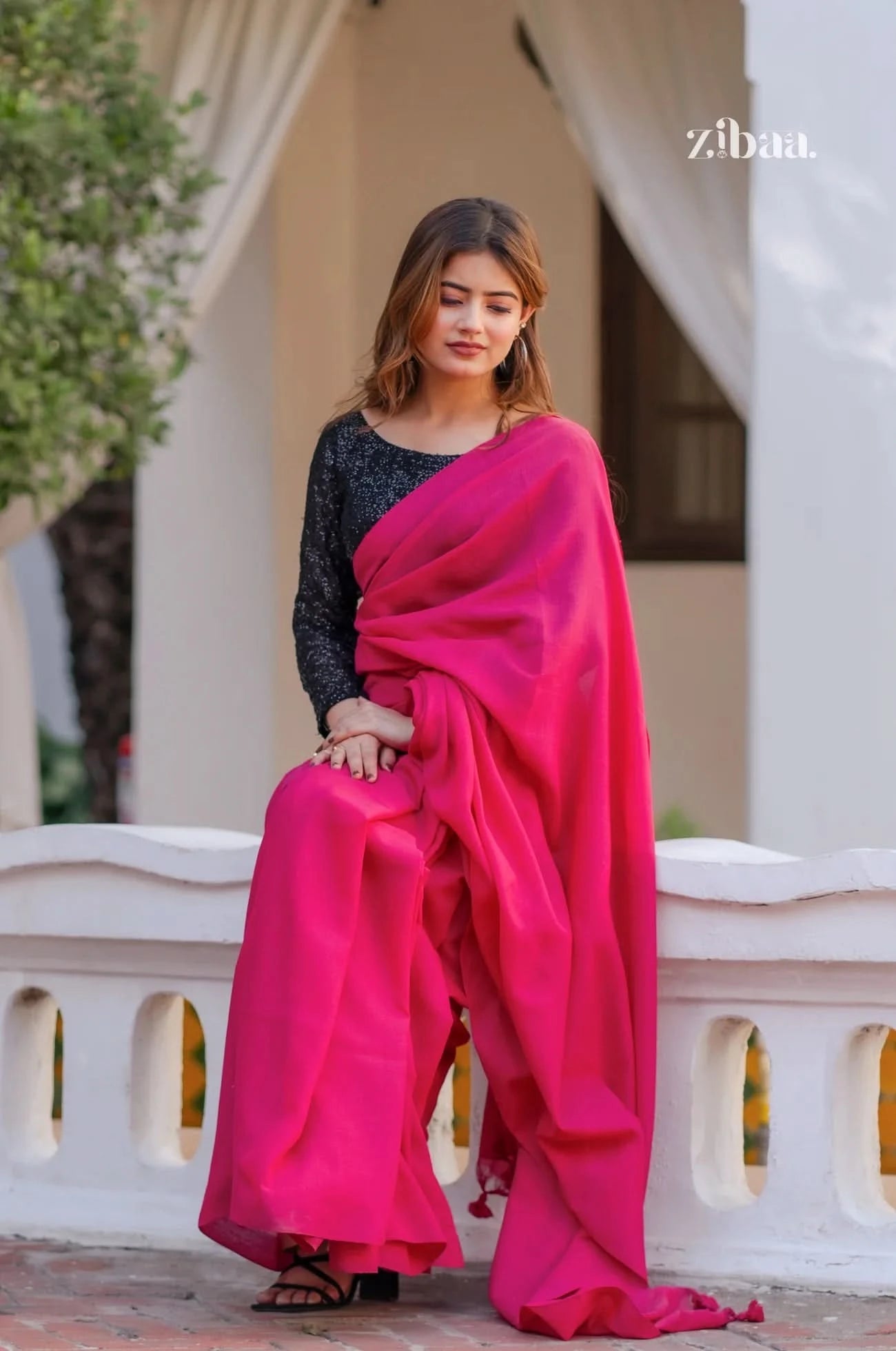 Woman in a bright pink saree sitting on a white railing with 'zibaa' branding.