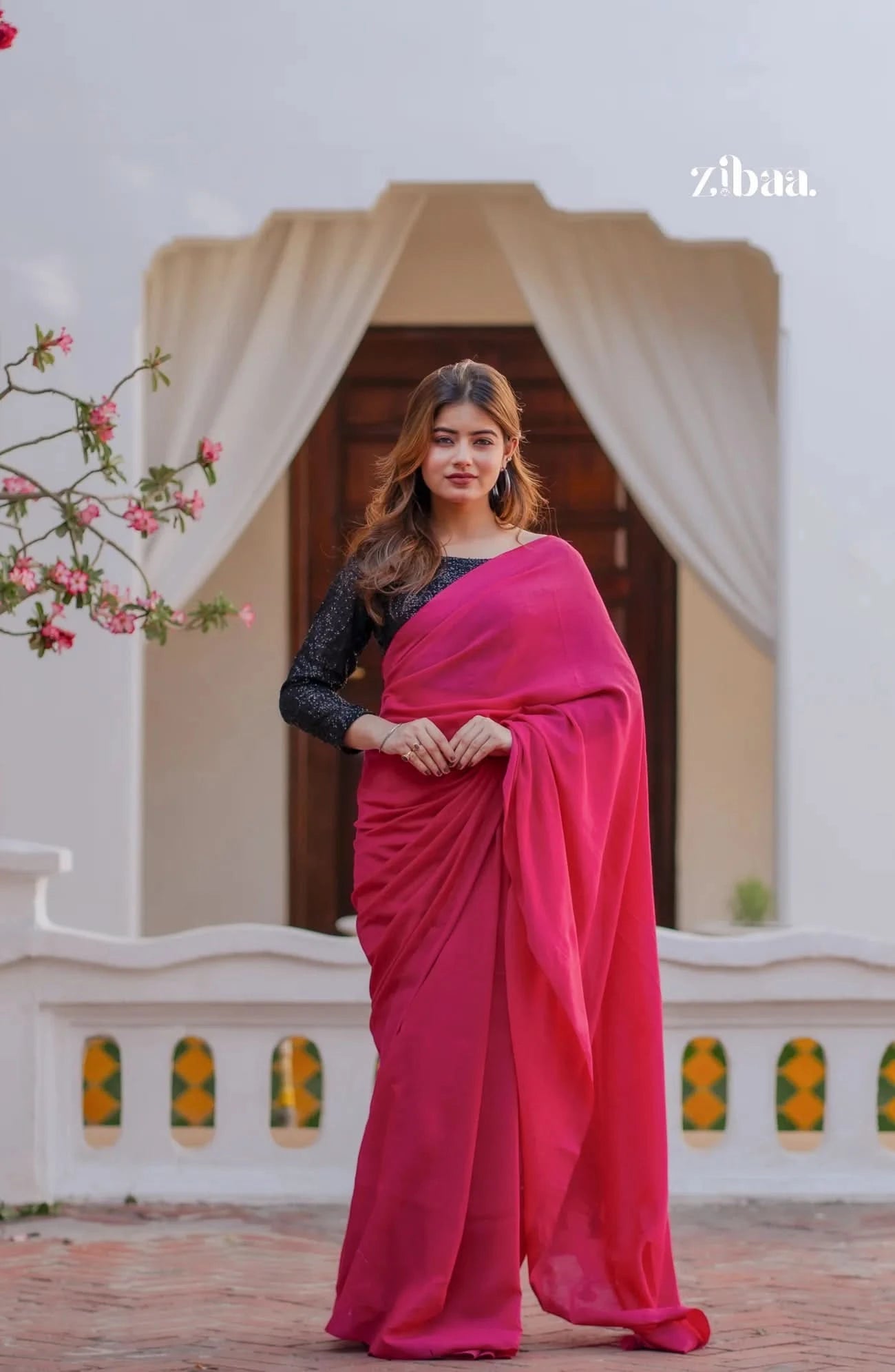 Woman in a pink saree standing in front of a decorative entrance with floral elements.
