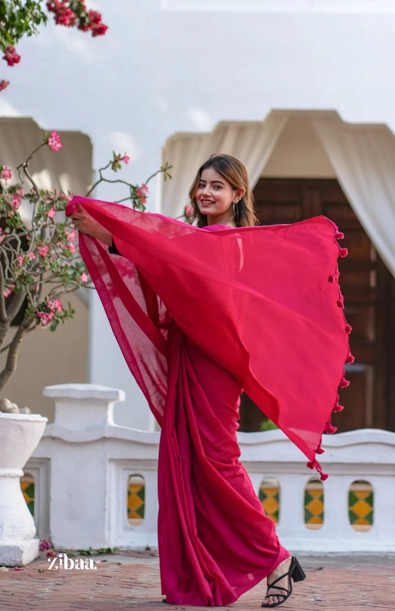 Woman in a red saree holding it up outdoors with floral decorations and a building in the background.