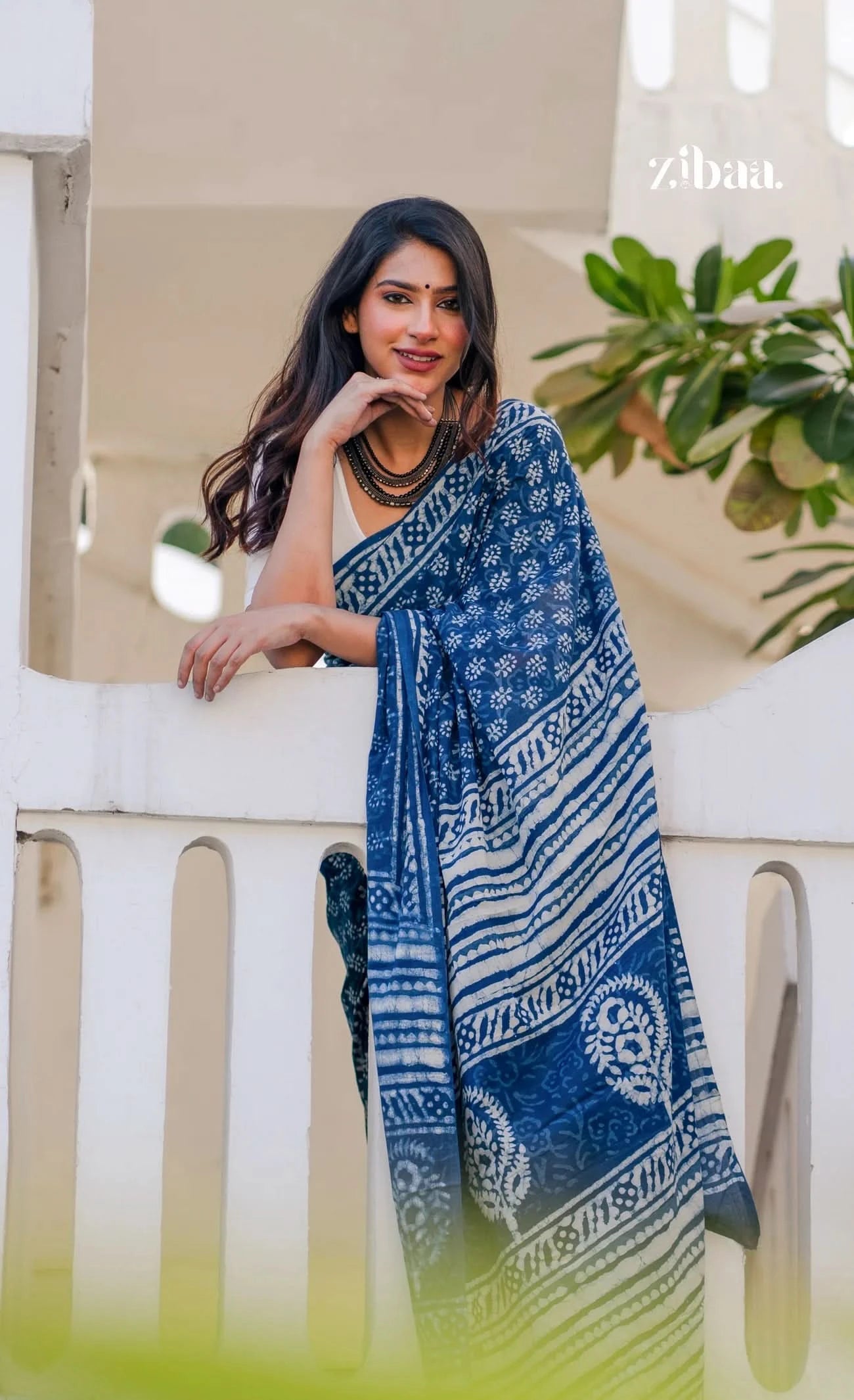 Woman wearing a blue and white patterned saree standing outdoors with plants in the background