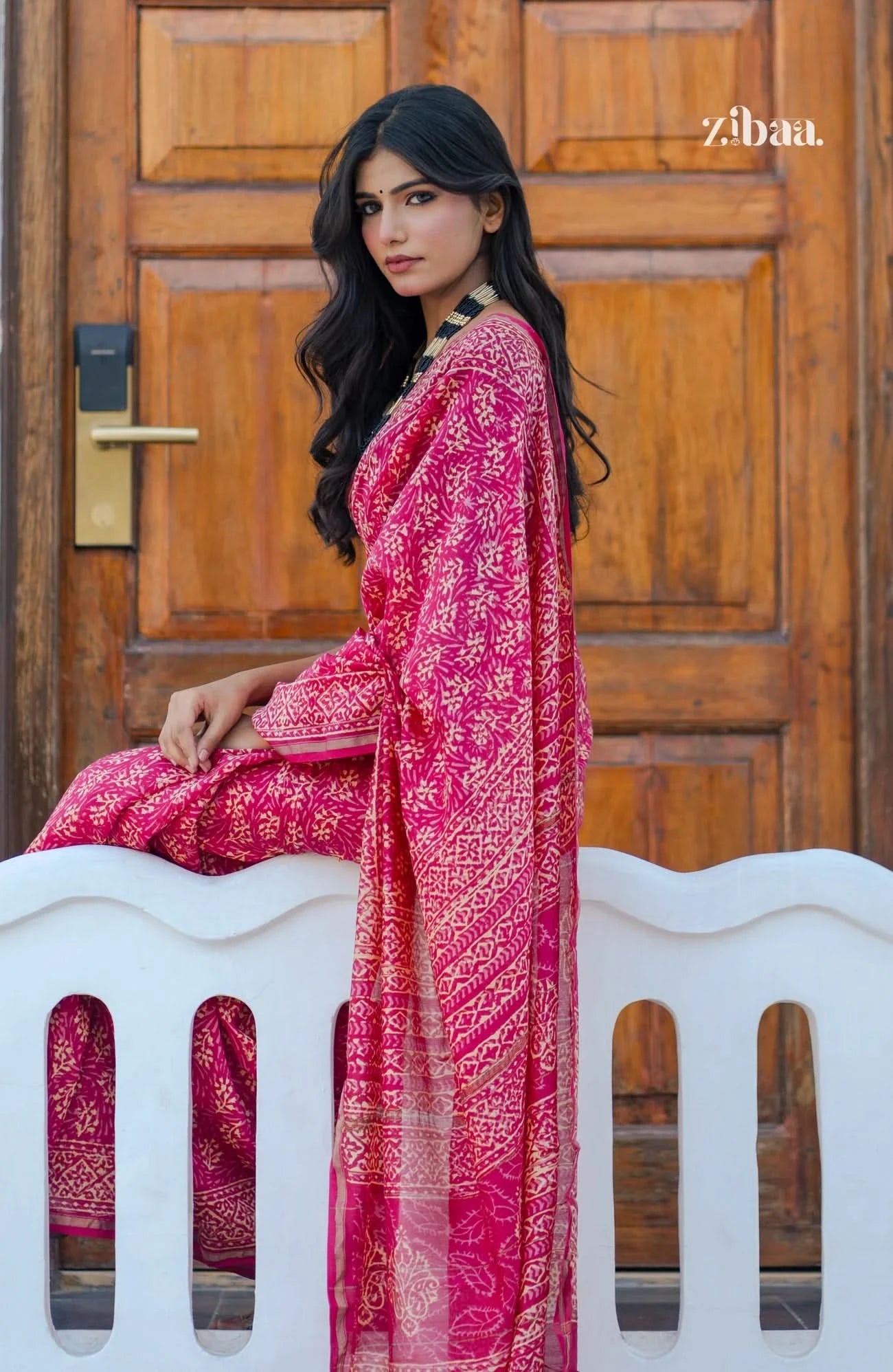 Woman in a pink saree sitting on a white bench with a wooden door in the background