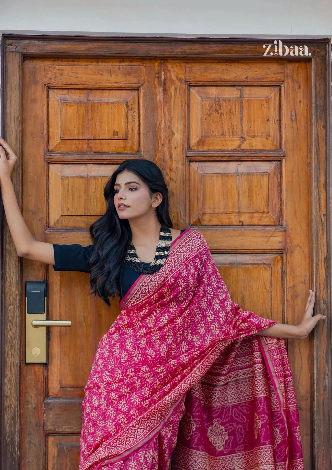 Woman in a pink saree standing in front of a wooden door with 'zibaa' branding.