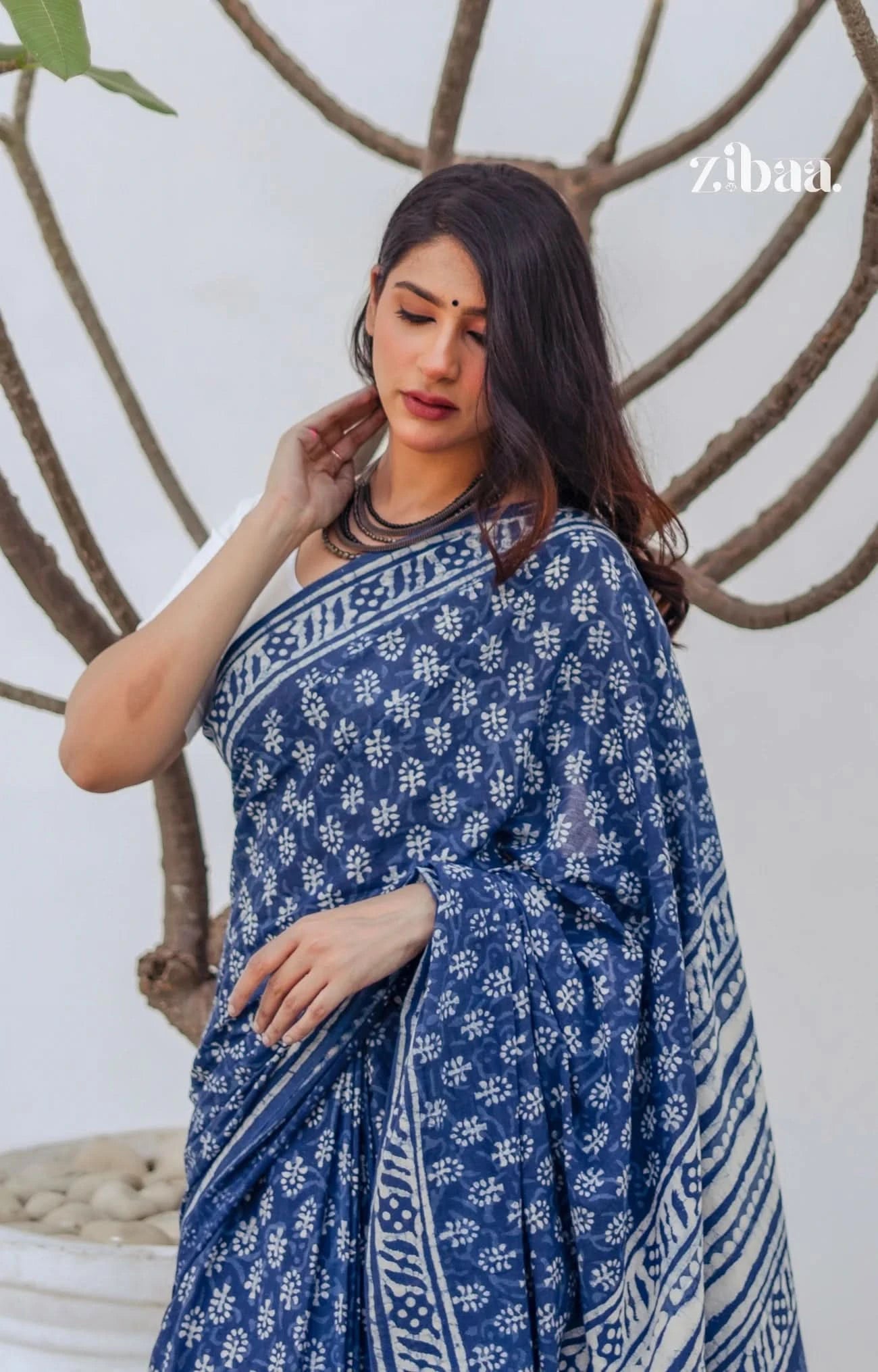 Woman wearing a blue saree with white patterns, standing against a neutral background with 'zibam' branding.