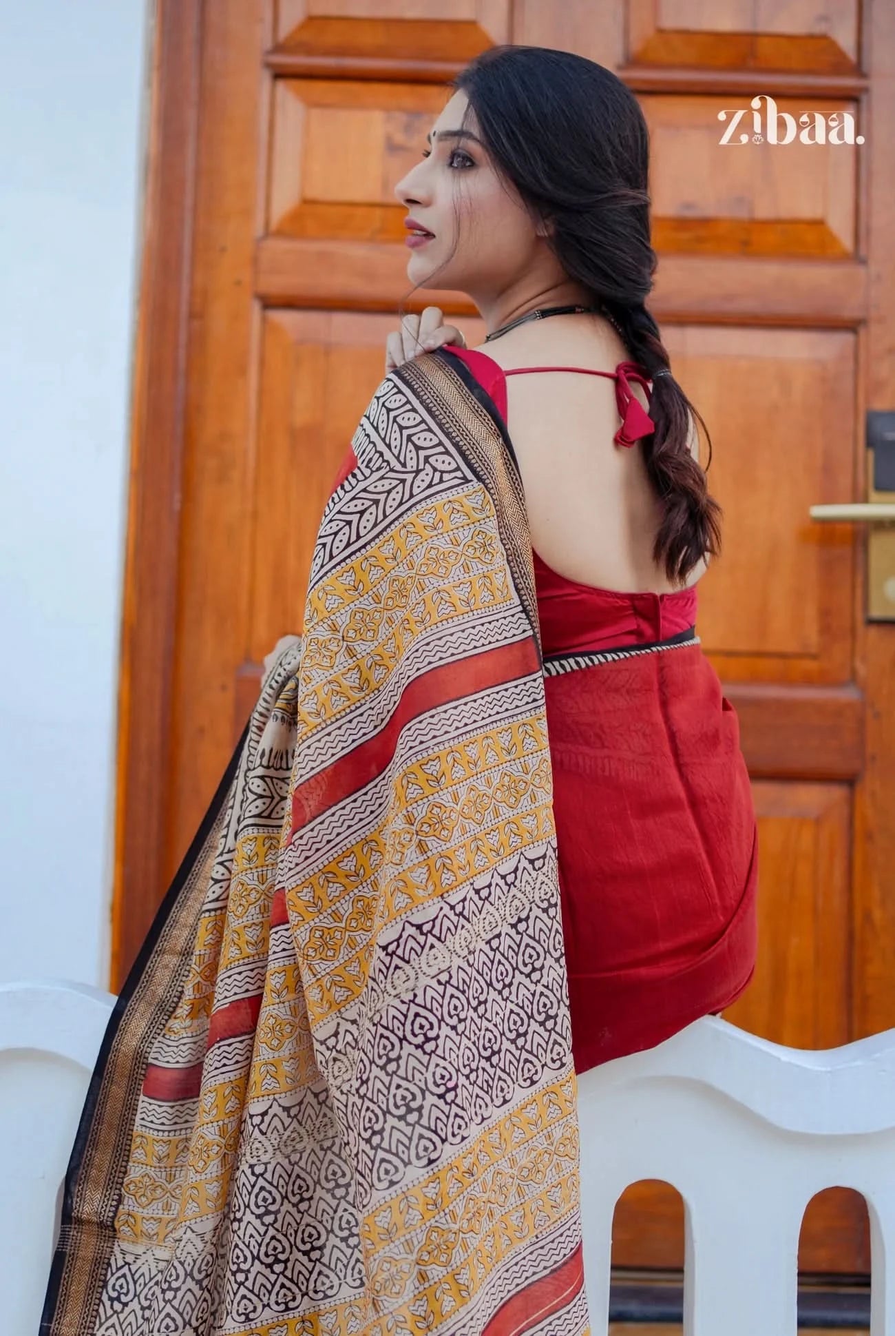 Woman wearing a red saree with a patterned shawl, standing in front of a wooden door.