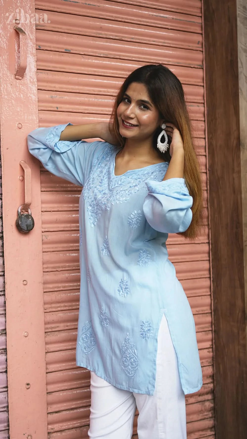 Woman in a light blue embroidered top and white pants standing against a pink shutter.