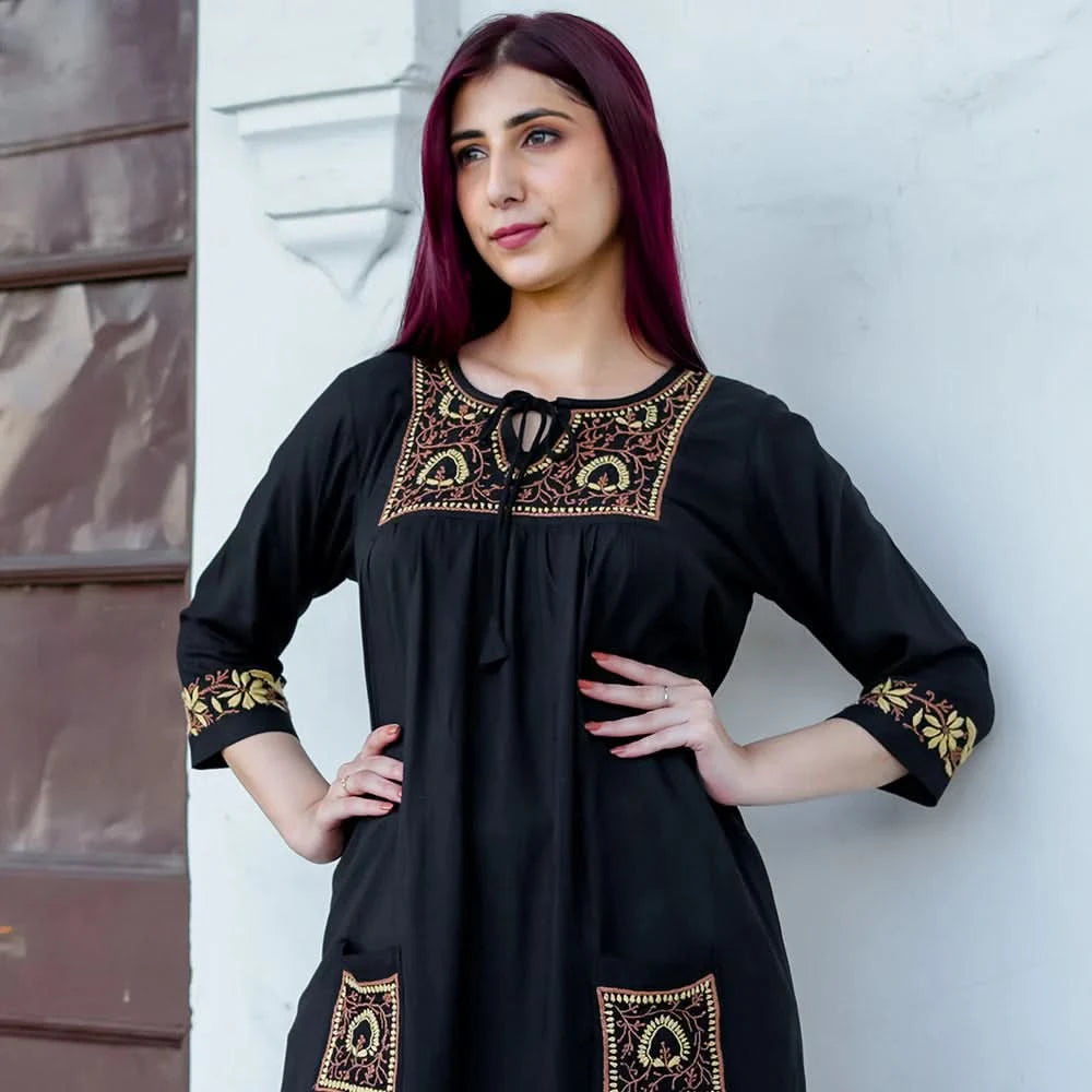 Woman wearing a black dress with gold embroidery against a white wall.