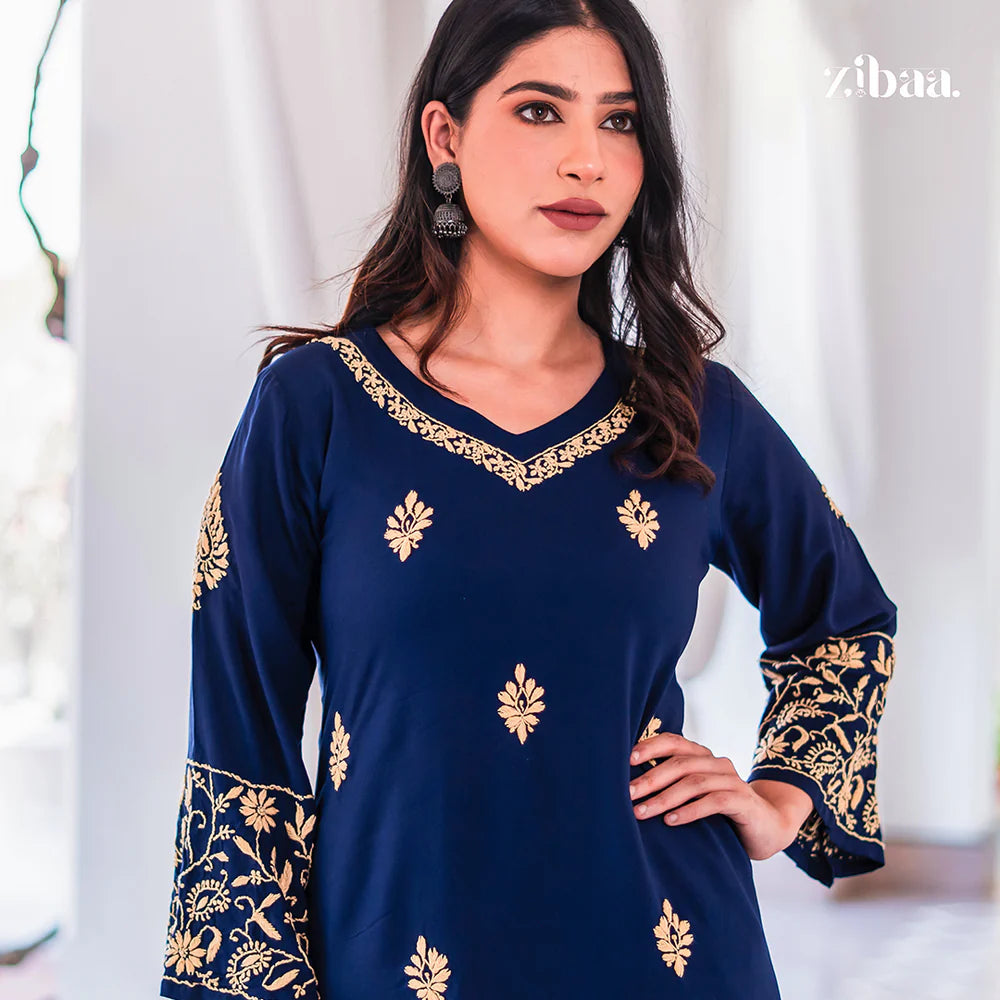Woman wearing a navy blue dress with gold embroidery against a white curtain background