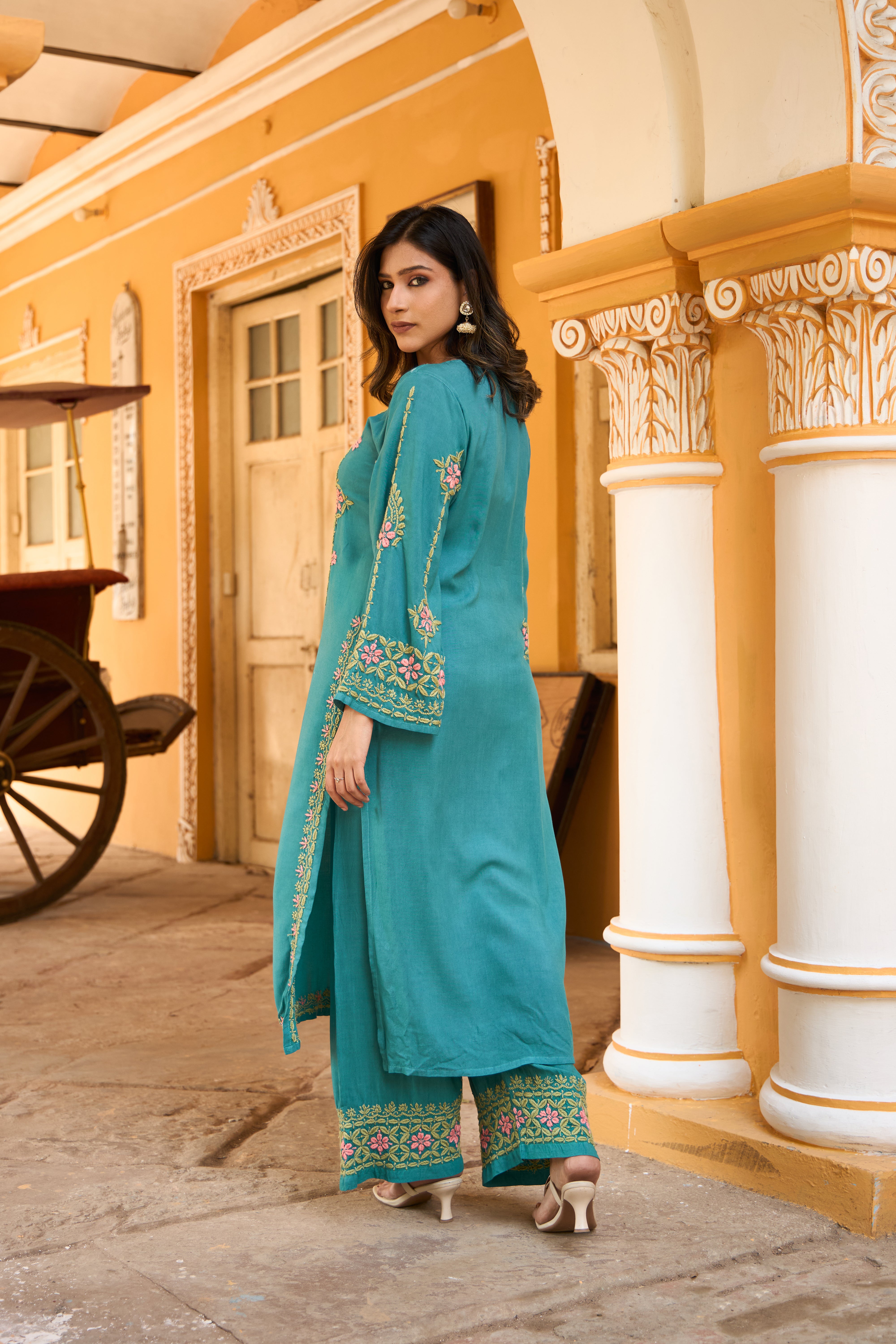 Woman in a teal traditional Chikankari kurta set standing in front of a yellow building with white columns.
