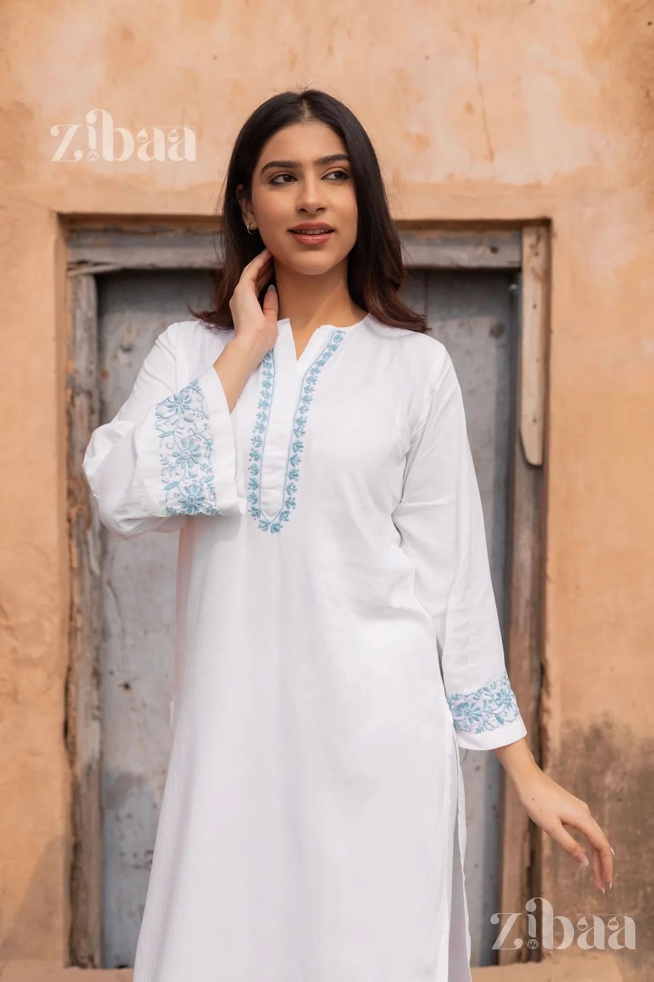 Woman wearing a white embroidered kurta standing against a rustic wall with 'zibaa' branding.