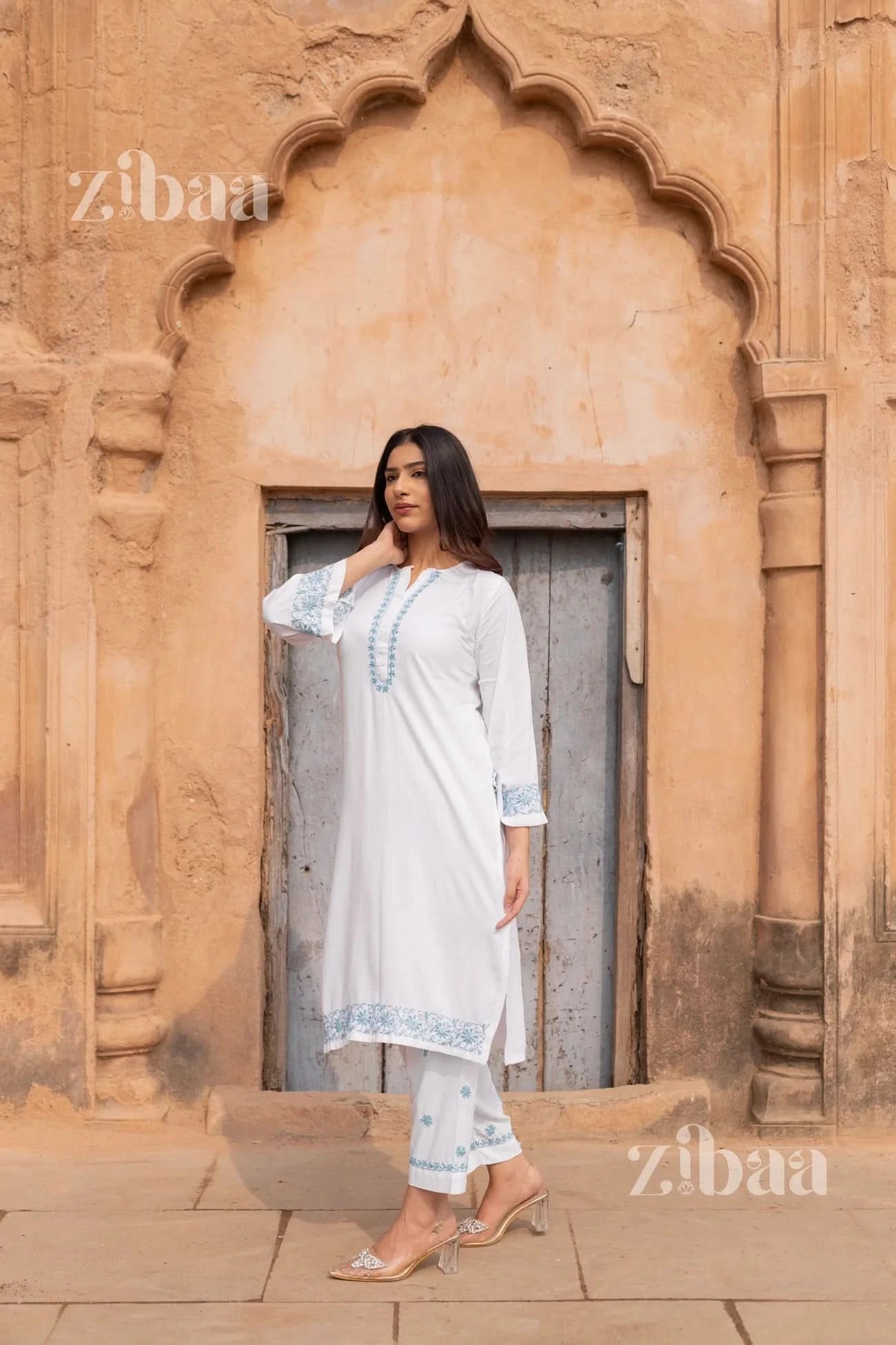 Woman in a white traditional outfit standing in front of an archway with 'zibaa' branding.