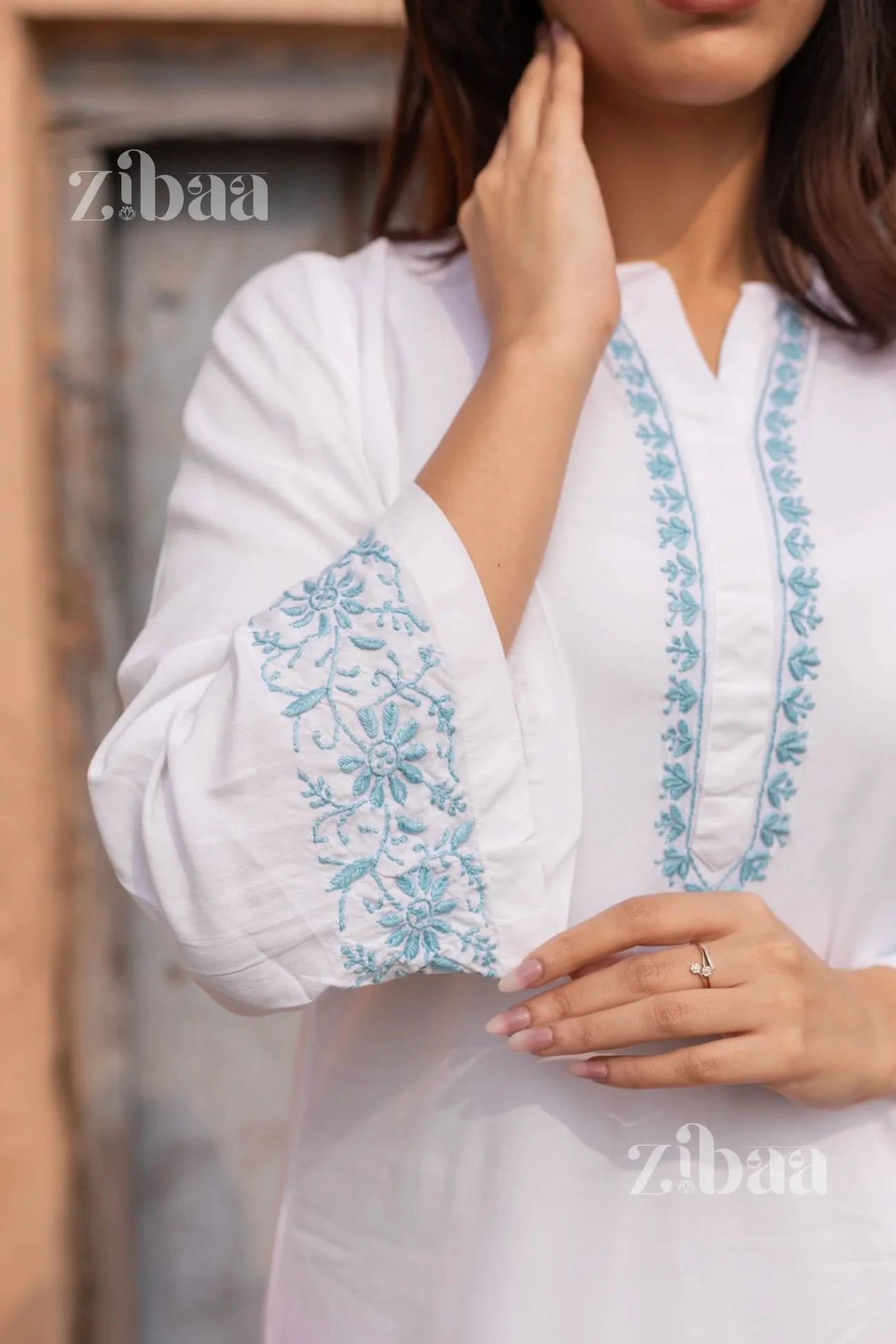 A woman wearing a white short kurti with white floral embroidery stands outdoors, touching her hair with one hand, gazing thoughtfully.