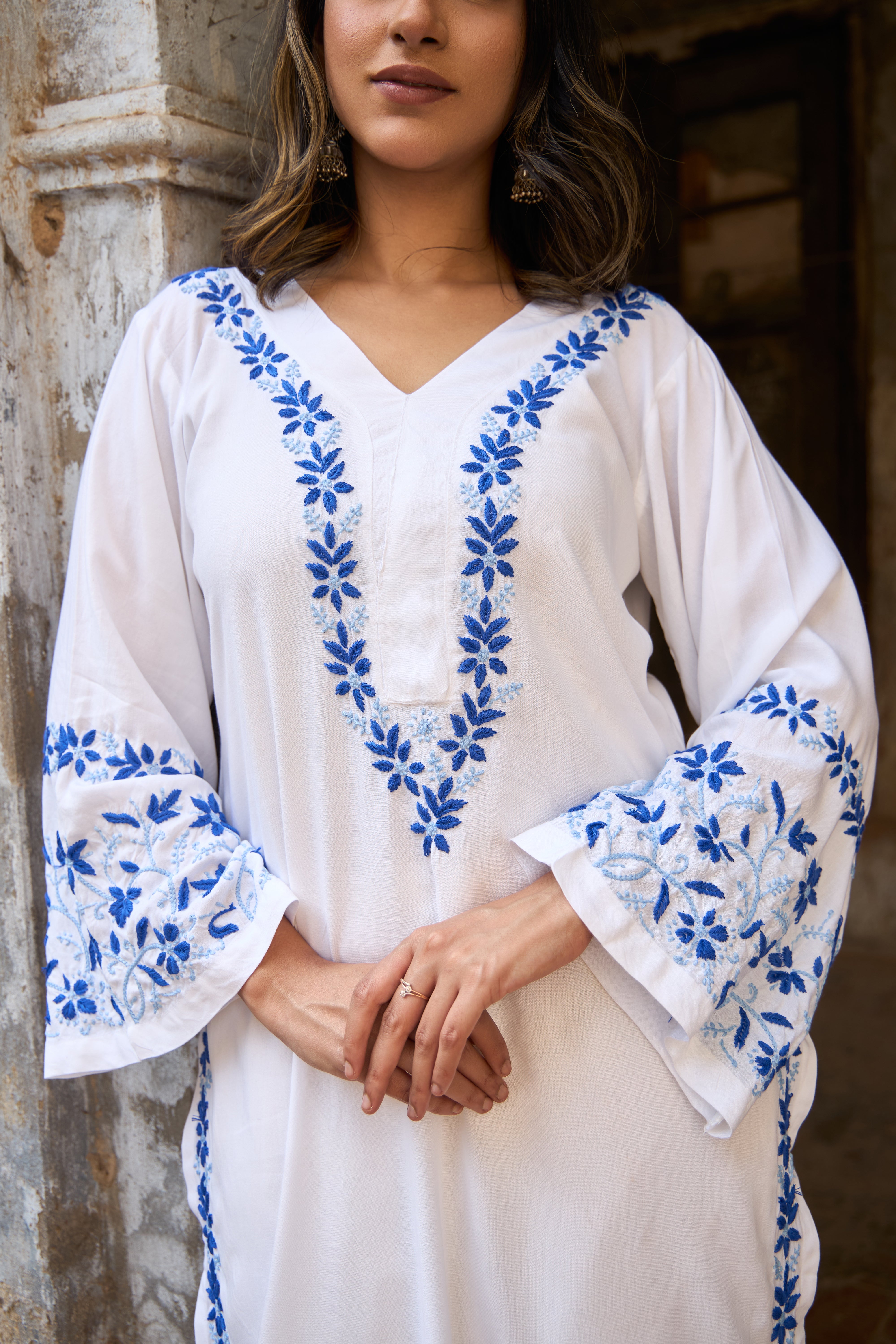 Woman wearing a White Chikankari Kurta with blue floral embroidery against a rustic stone wall.