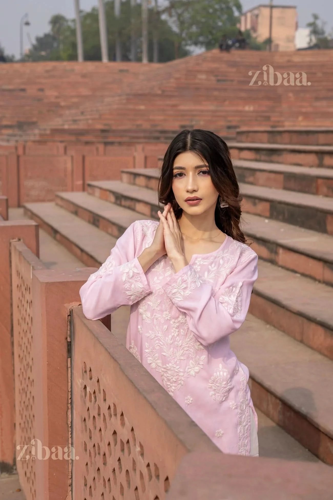 Model wearing a pastel pink Lucknowi Kurti with white embroidery, posing against a rustic backdrop of steps and traditional architecture