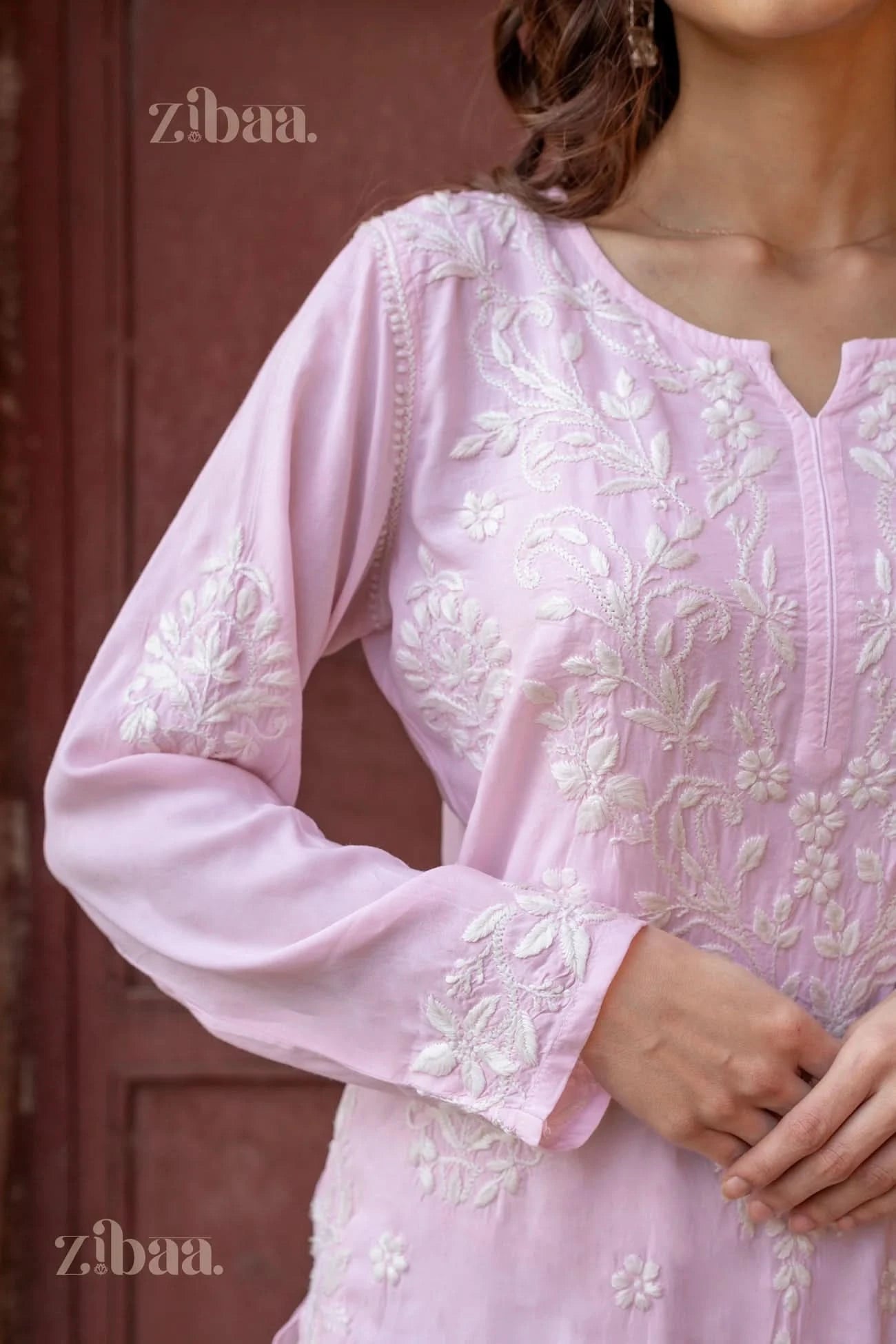 Model wearing a pastel pink Chikankari Kurti with white embroidery, paired with white pants, posing gracefully in a traditional setting.